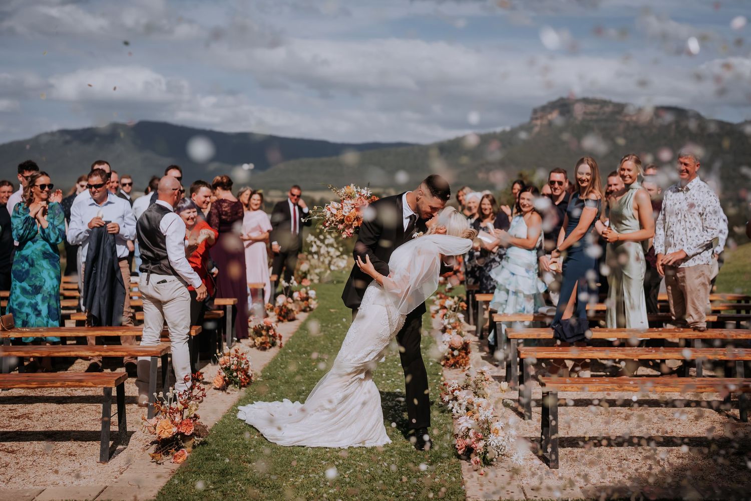Bride and groom share a romantic kiss at outdoor wedding ceremony with floral aisle and mountain backdrop.