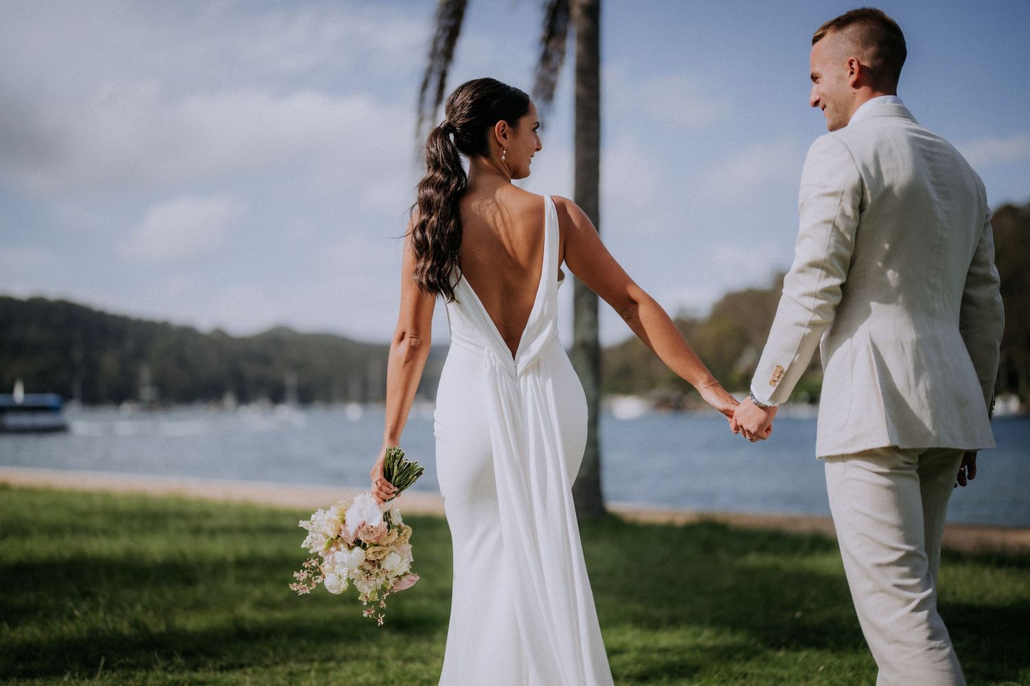 Elegant waterfront wedding portrait showing bride in backless dress holding bouquet.