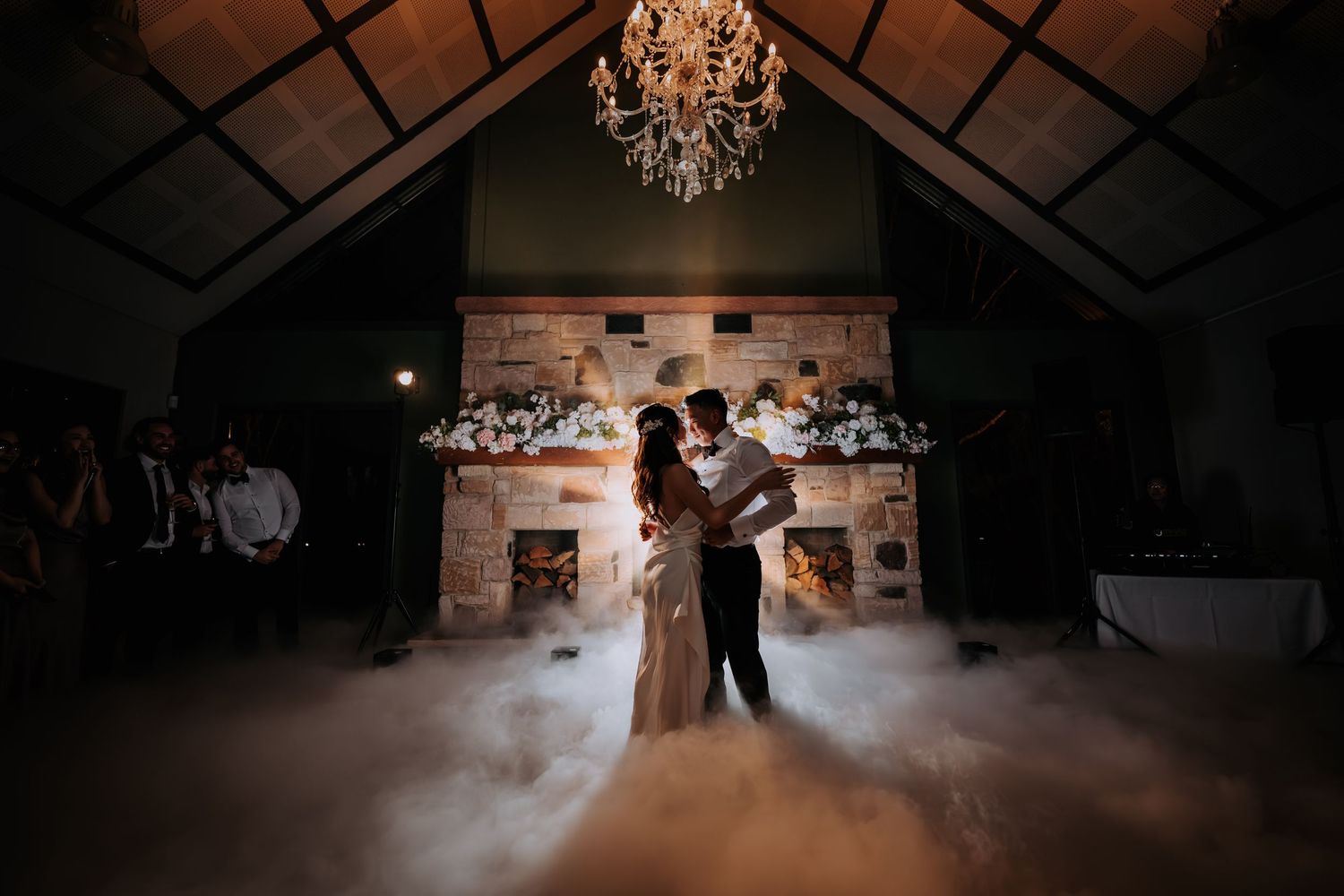 Couple shares first dance surrounded by dry ice fog under chandelier with stone fireplace backdrop at elegant wedding reception.