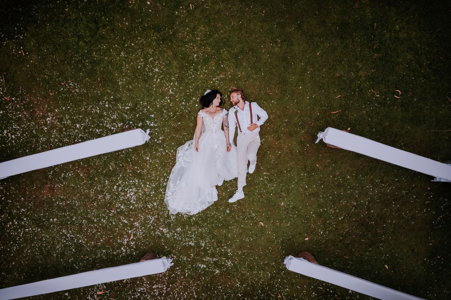 Aerial view of bride and groom lying on green grass surrounded by white ribbon decorations at outdoor wedding.