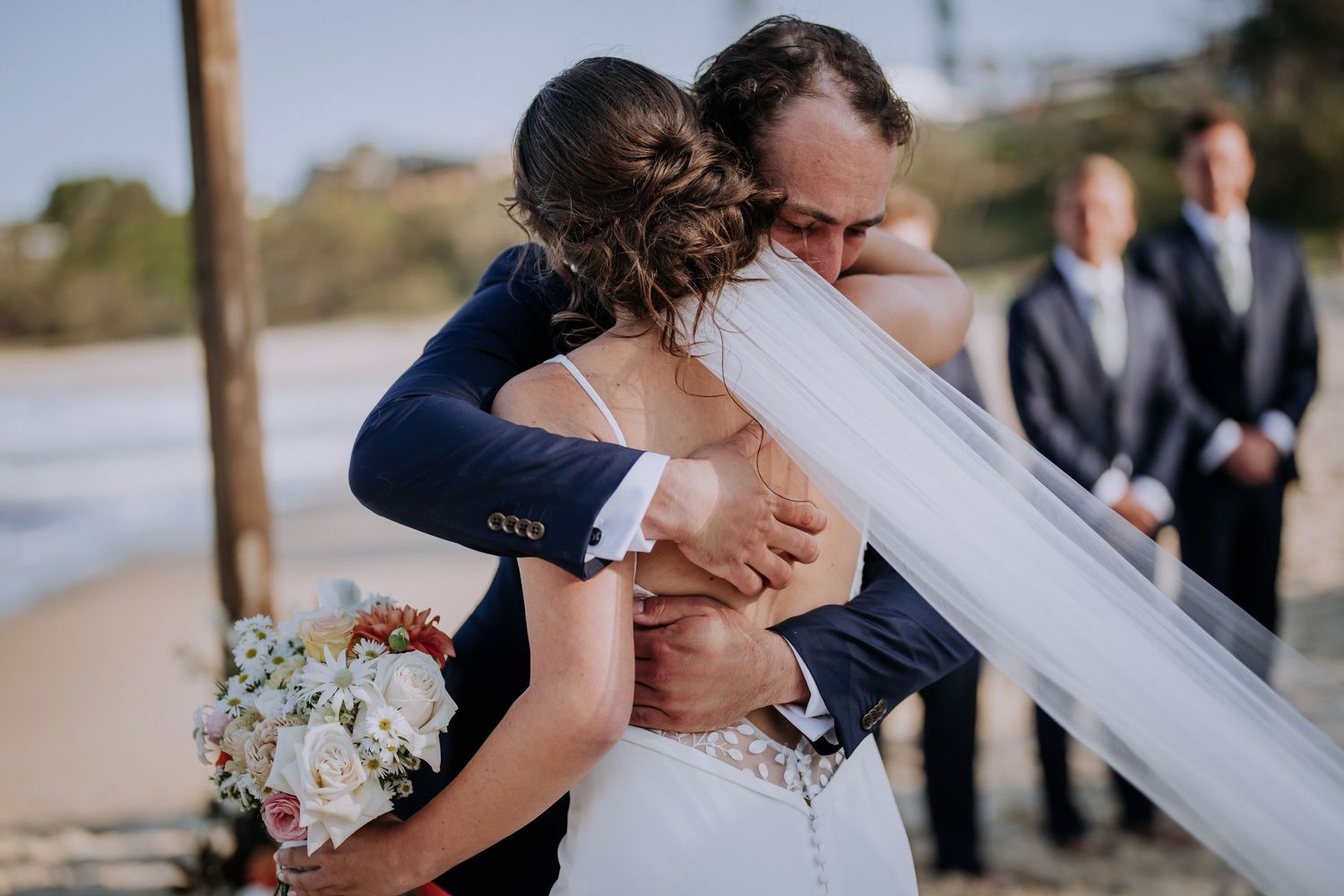 Emotional beachside wedding ceremony with couple sharing first kiss as guests look on.