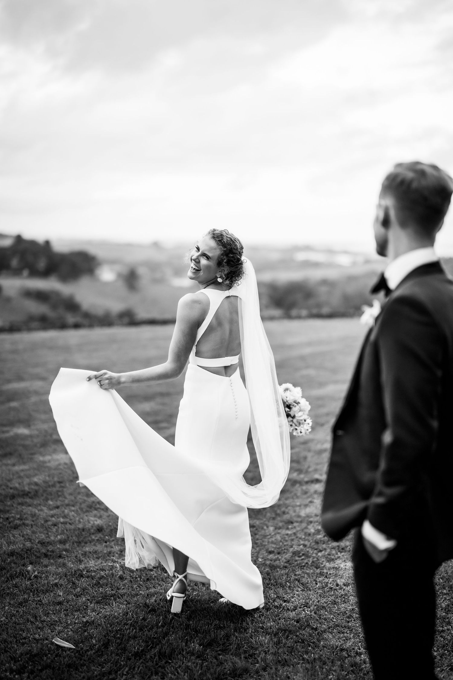 Elegant bride in backless dress twirls on hillside with flowing veil.