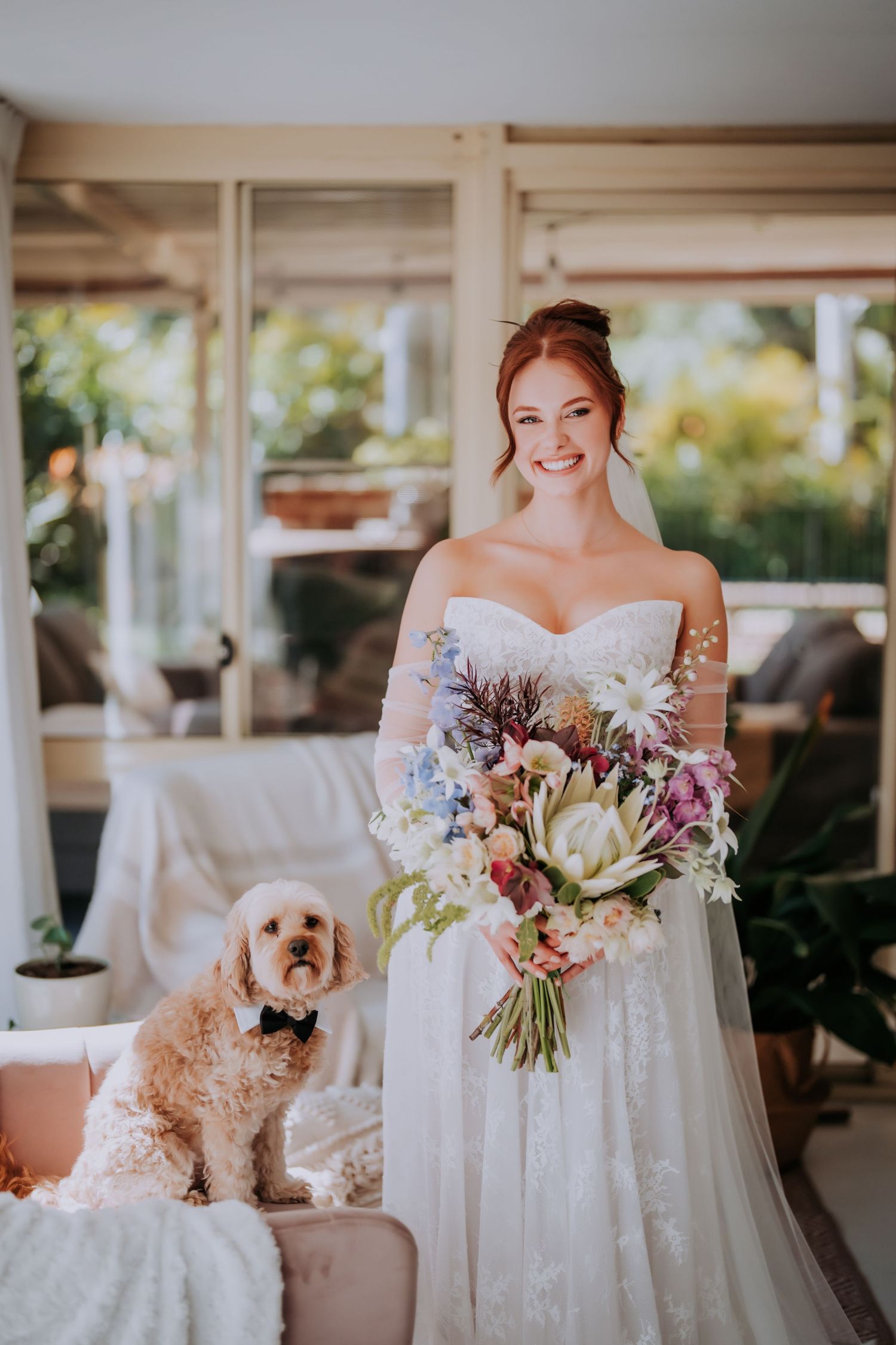 Smiling bride in white gown holds colorful bouquet beside a fluffy dog wearing a bow tie indoors.