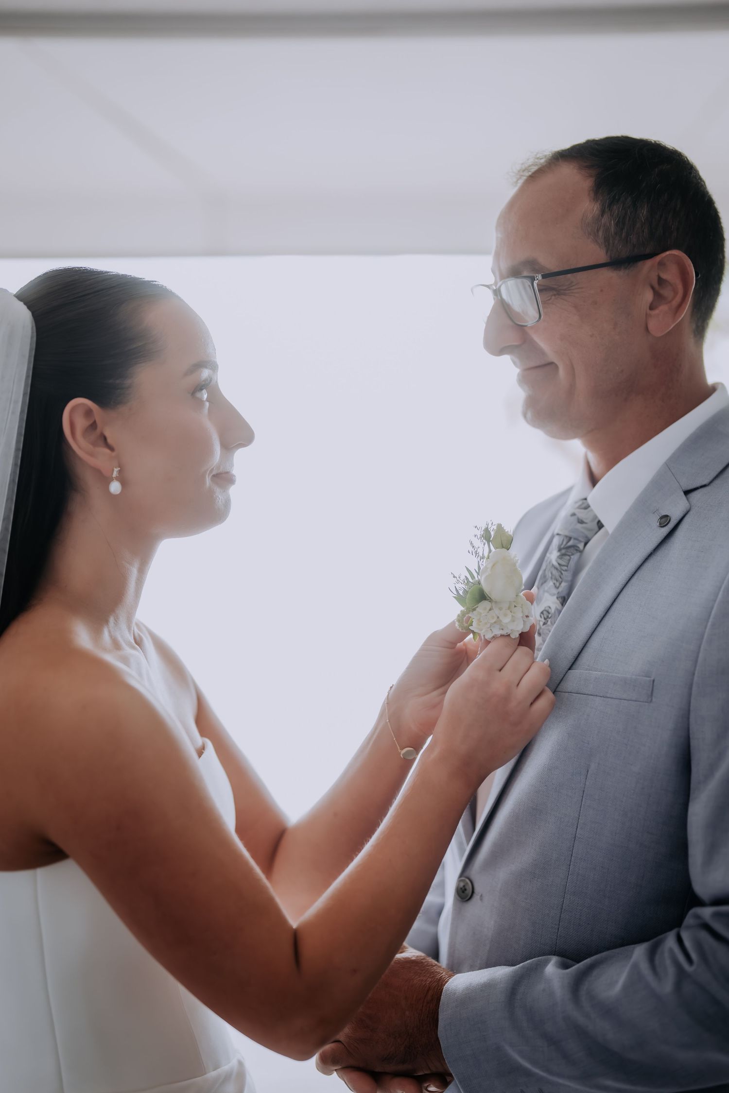 Bride in white dress pins a boutonniere on groom in gray suit, sharing a tender moment before wedding ceremony.