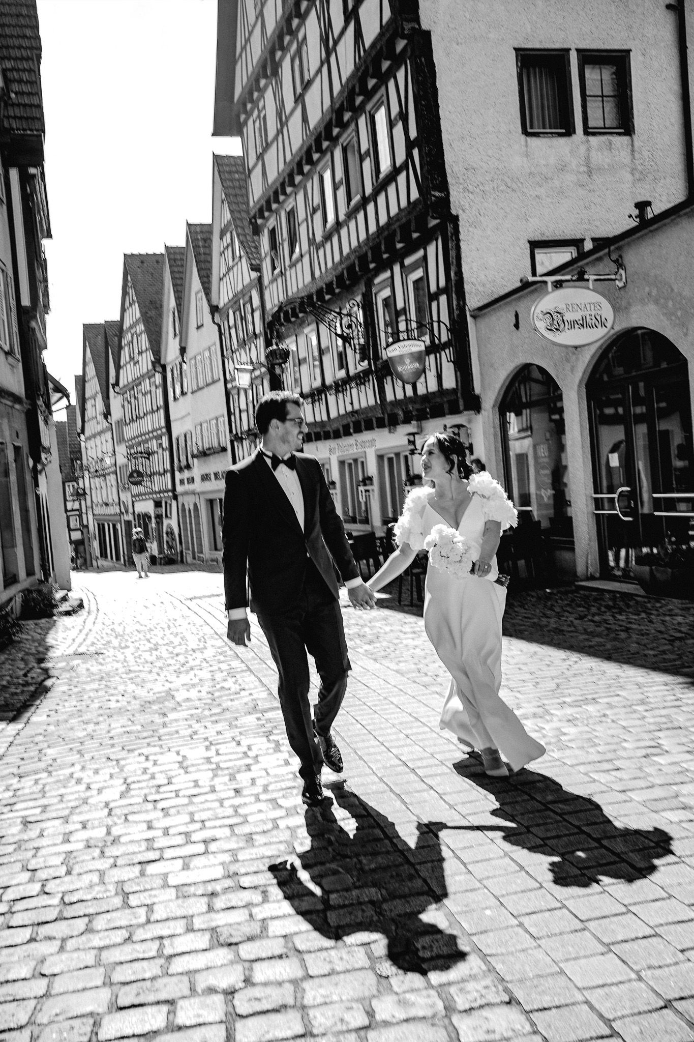 A couple in wedding attire walks along a cobblestone street lined with traditional half-timbered buildings in Germany.