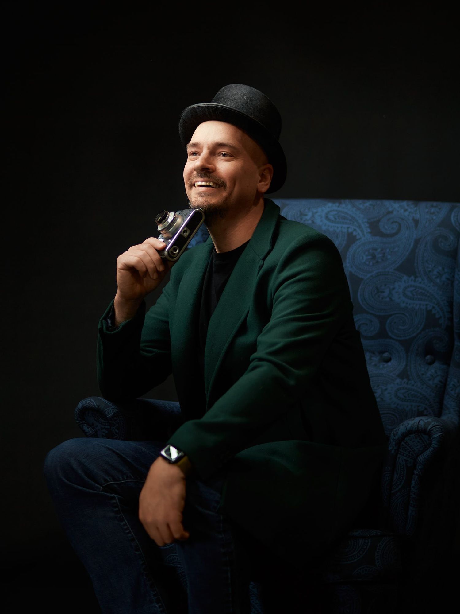 Photographer Alex Kalimat sitting in a wingback chair wearing a top hat and velvet jacket.