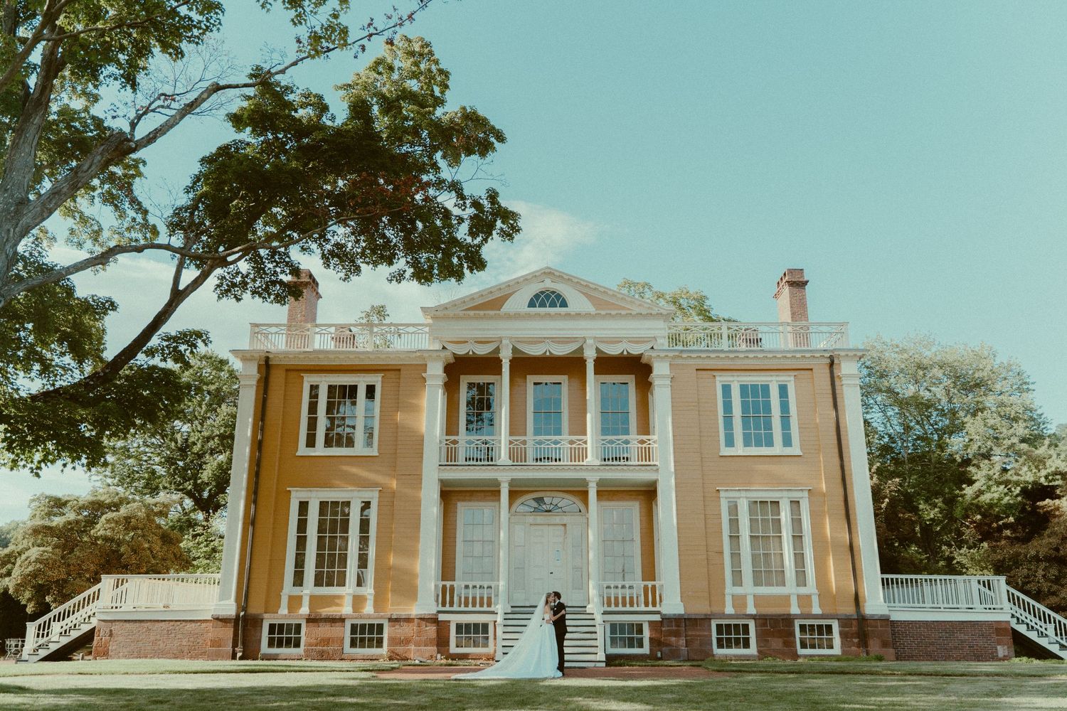 Historic peach-colored mansion with grand columns and symmetrical staircases set against a soft teal sky.