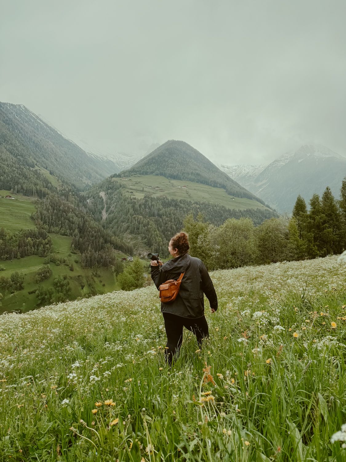 Person walking through a wildflower meadow with mountain peaks and cloudy skies in the background.