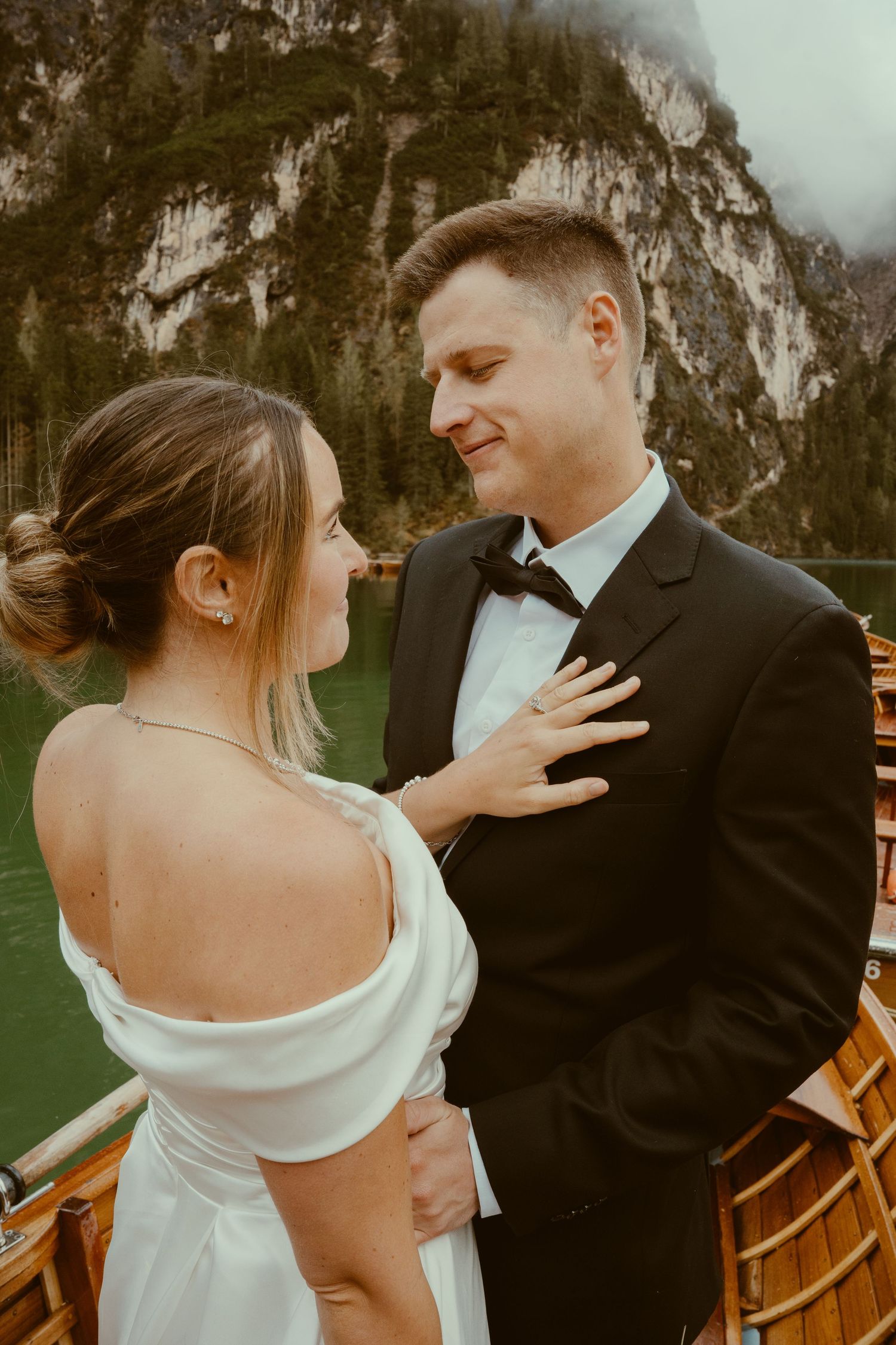 A couple shares a romantic moment on a wooden boat with dramatic mountain cliffs in the background.