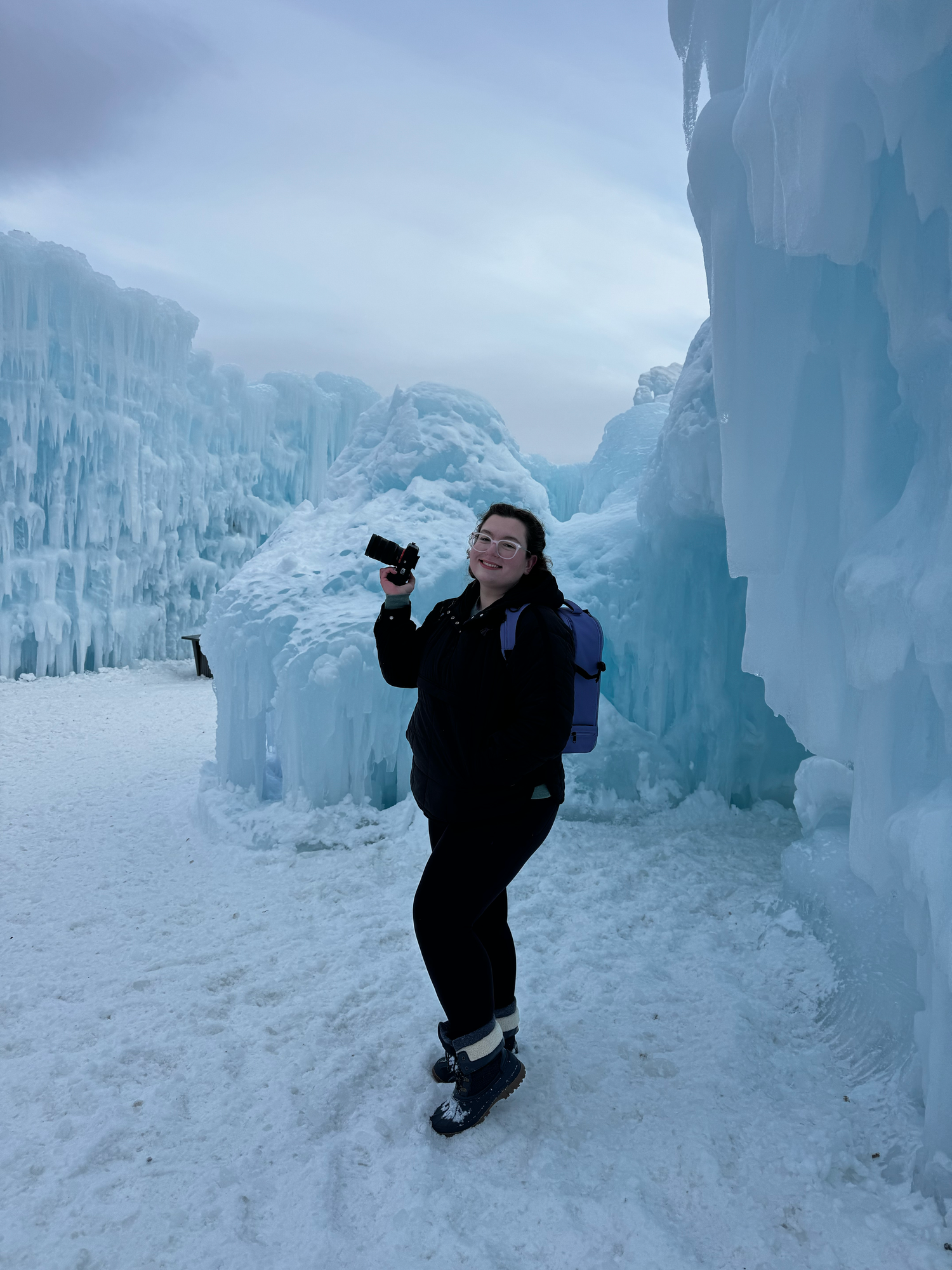 Someone poses in a winter outfit against towering ice formations at a frozen attraction in snowy conditions.