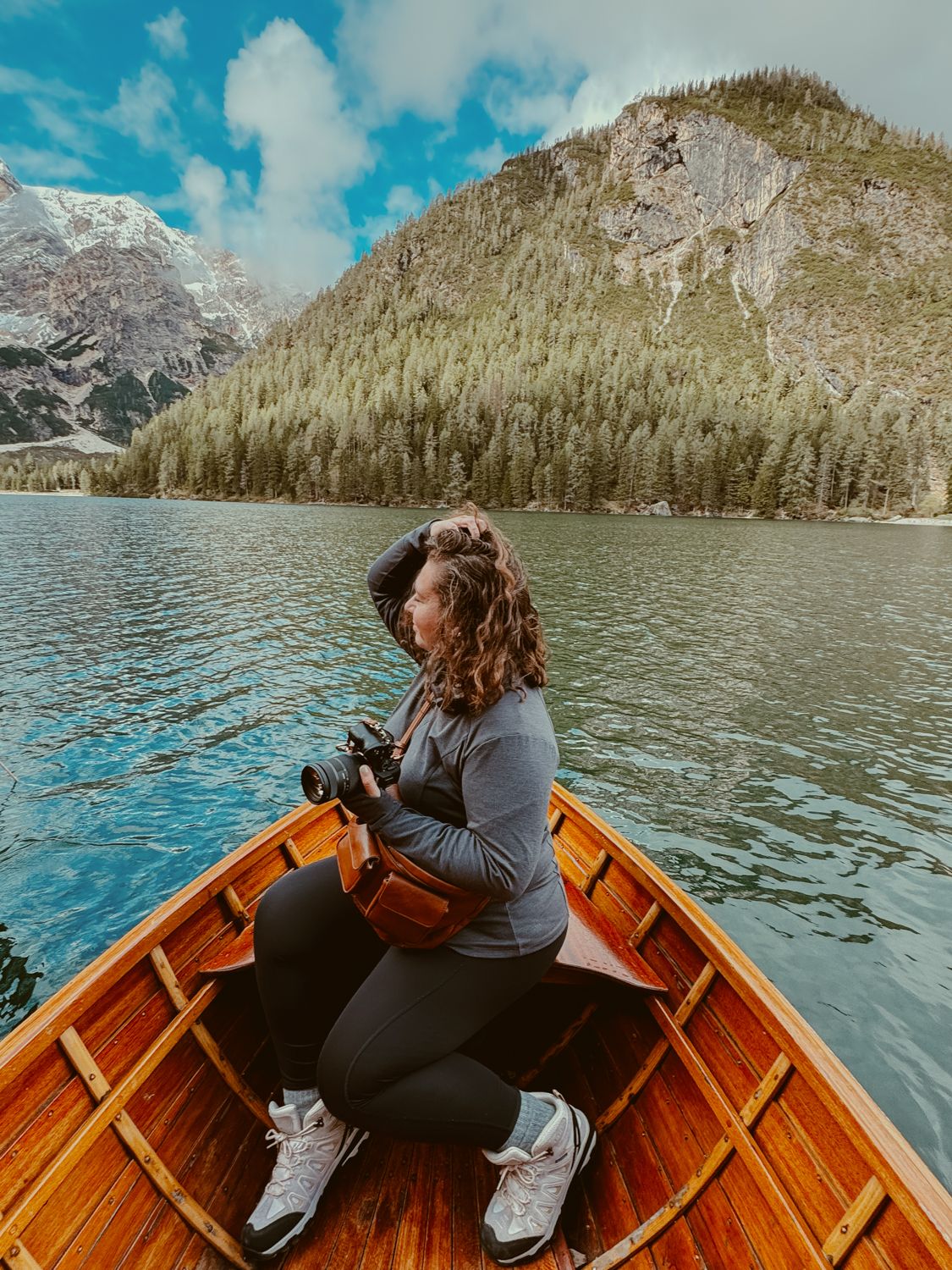 Someone sits in a wooden boat on a mountain lake surrounded by evergreen forests and snow-capped peaks on a sunny day.