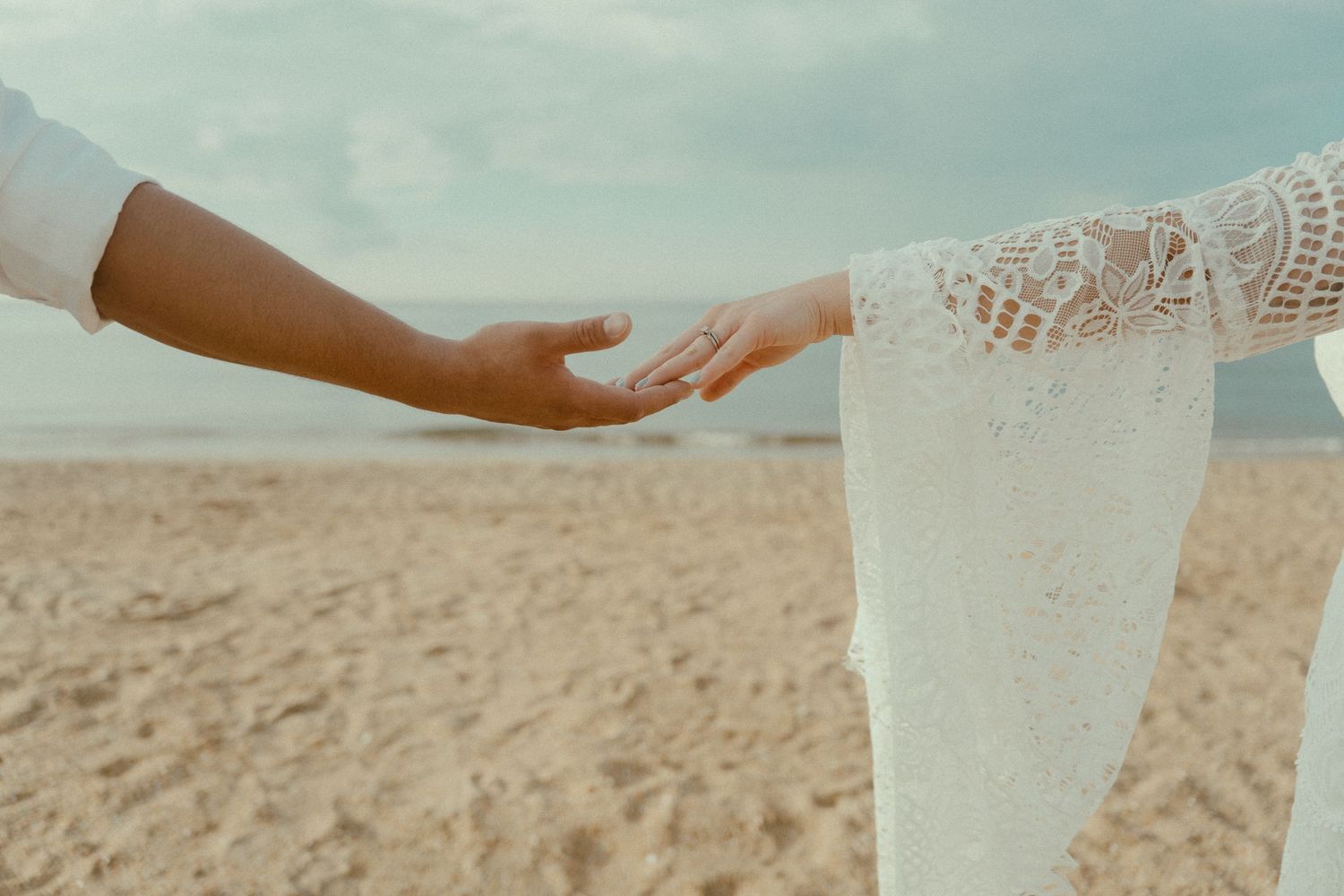 Close-up of two hands exchanging a white lace-trimmed handkerchief against a beige beach background.
