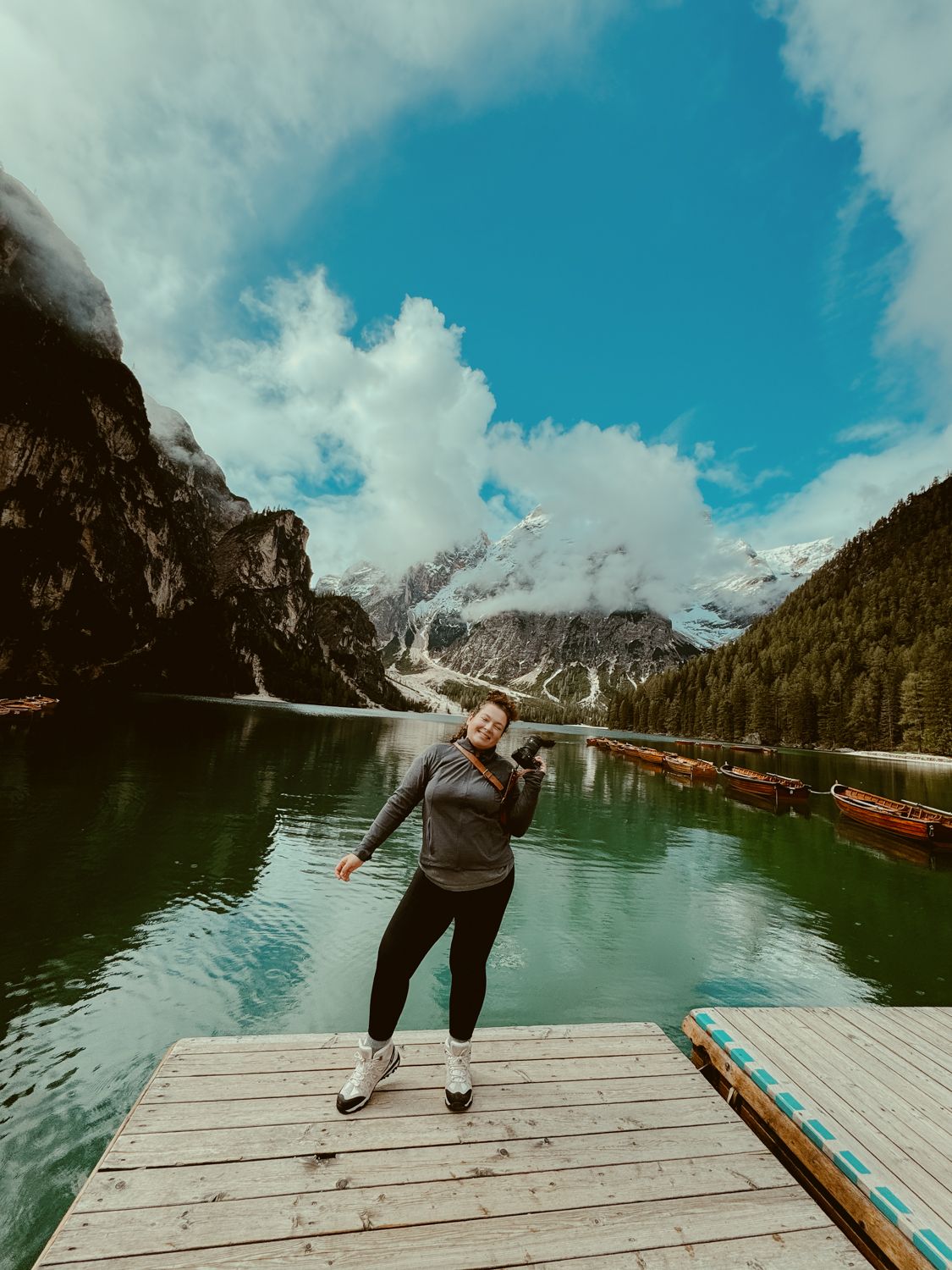 Person posing playfully on a wooden dock at a stunning alpine lake surrounded by snow-capped mountains and forest.