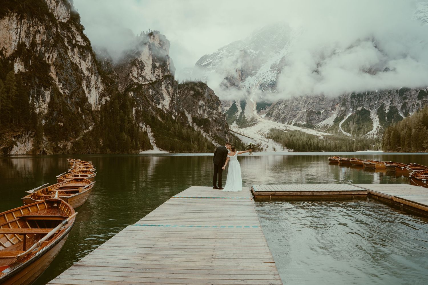 Wooden boats docked at a scenic mountain lake with snow-capped peaks and a long pier stretching into misty waters.