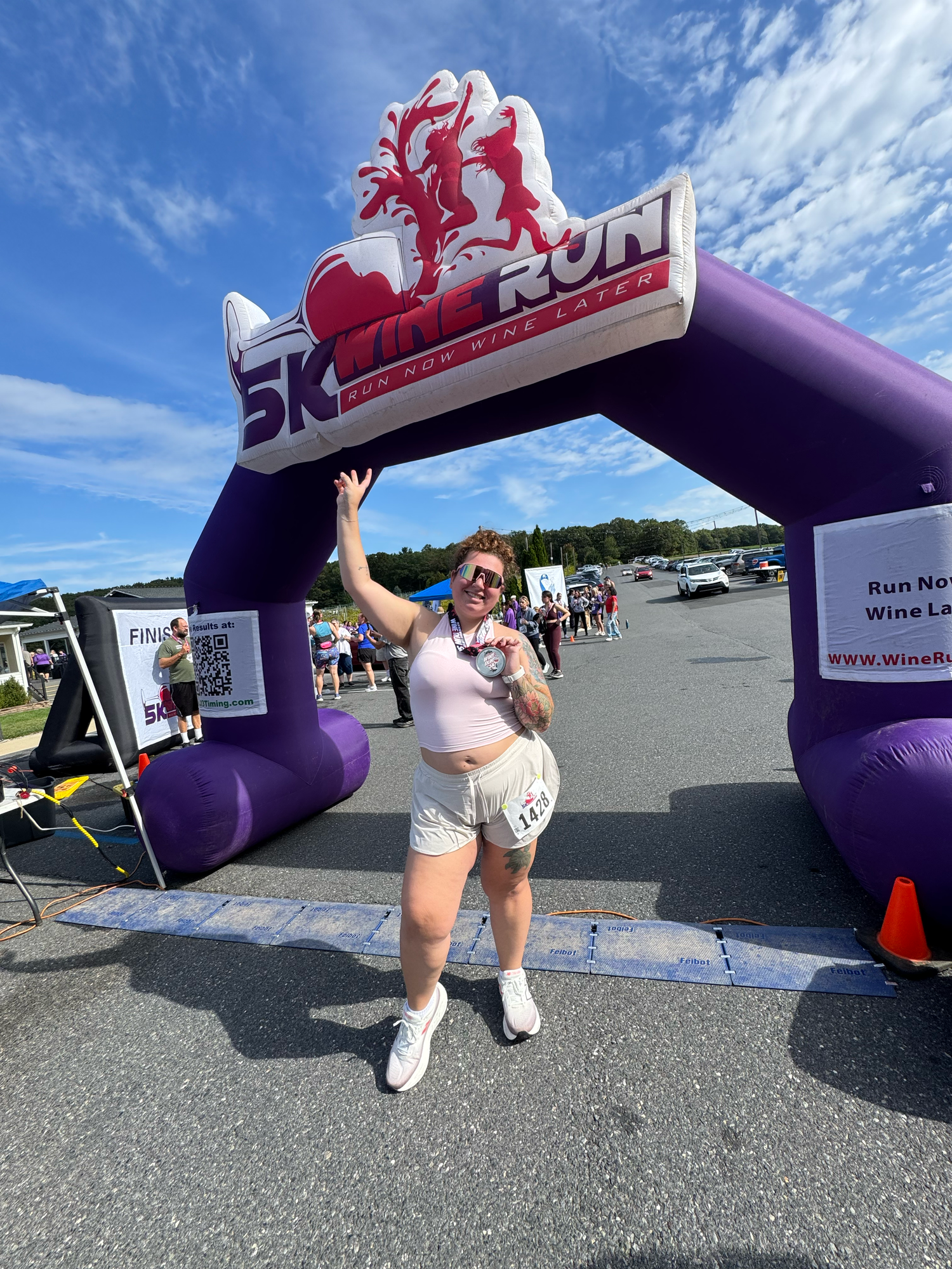 A participant poses enthusiastically under a purple inflatable 5K race arch against a bright blue sky.