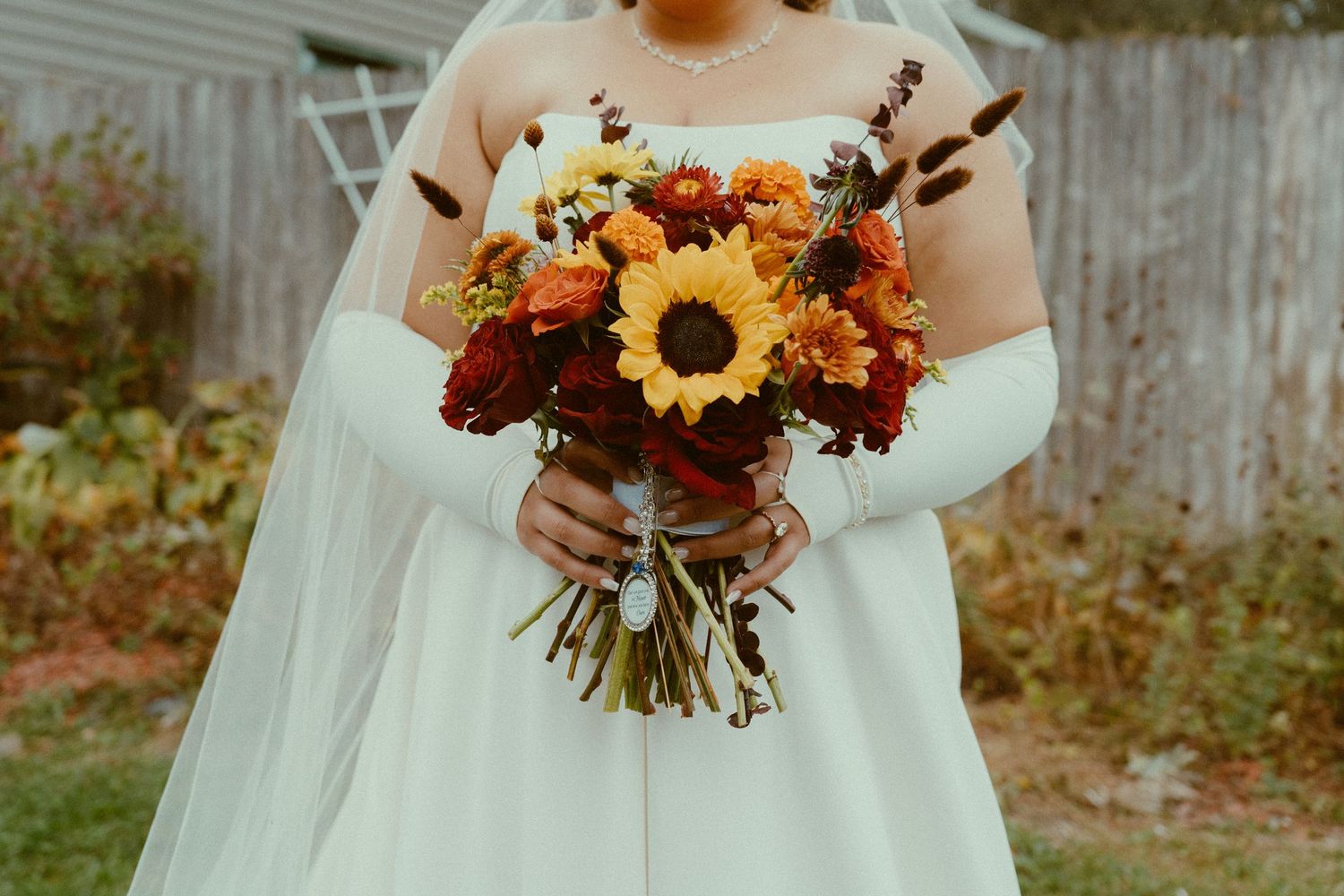 A beautiful autumn wedding bouquet featuring vibrant sunflowers, orange roses and rustic fall foliage against a wooden fence.