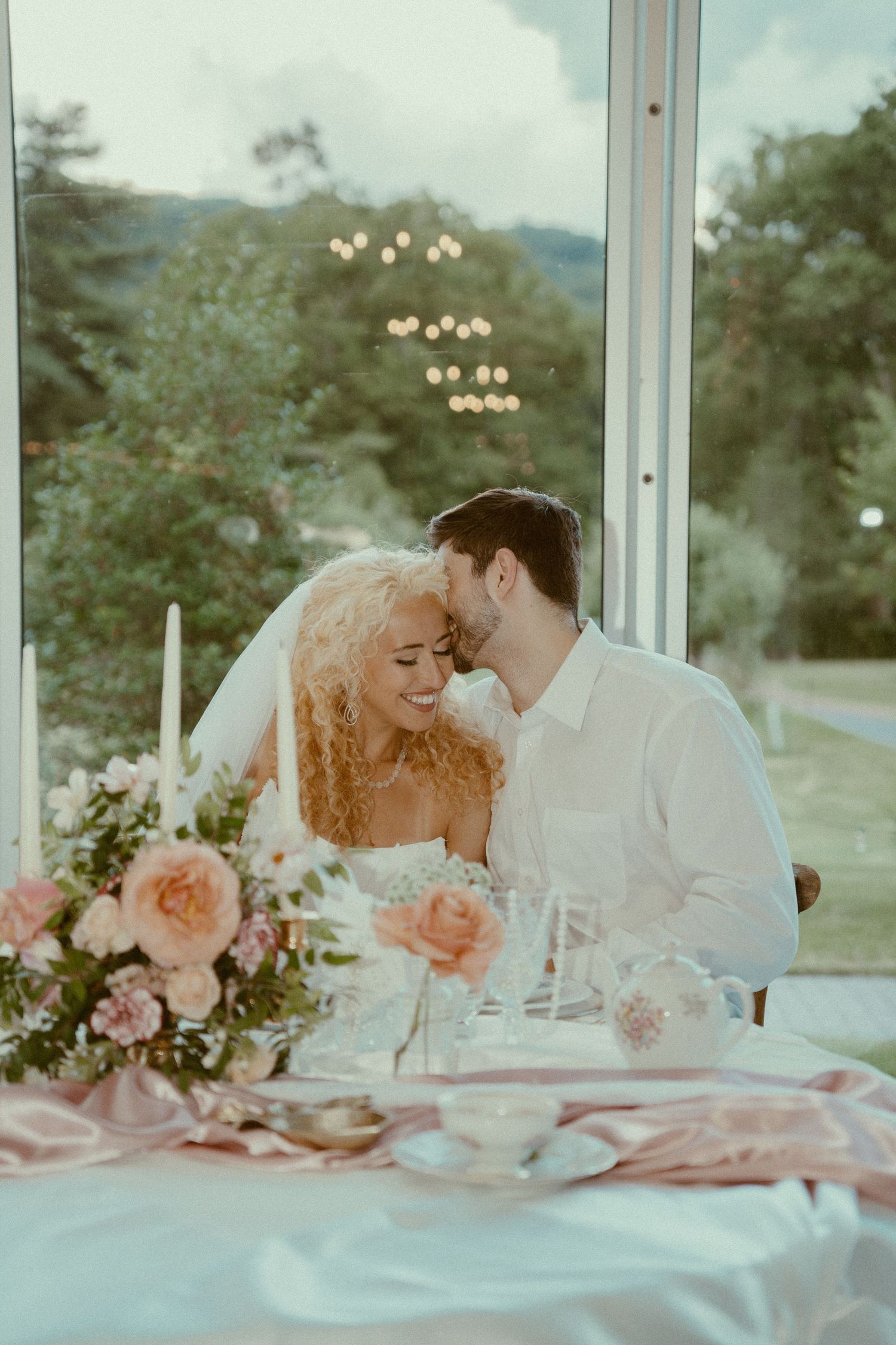 A romantic wedding table setting with pink flowers and soft lighting in a glass-enclosed venue.