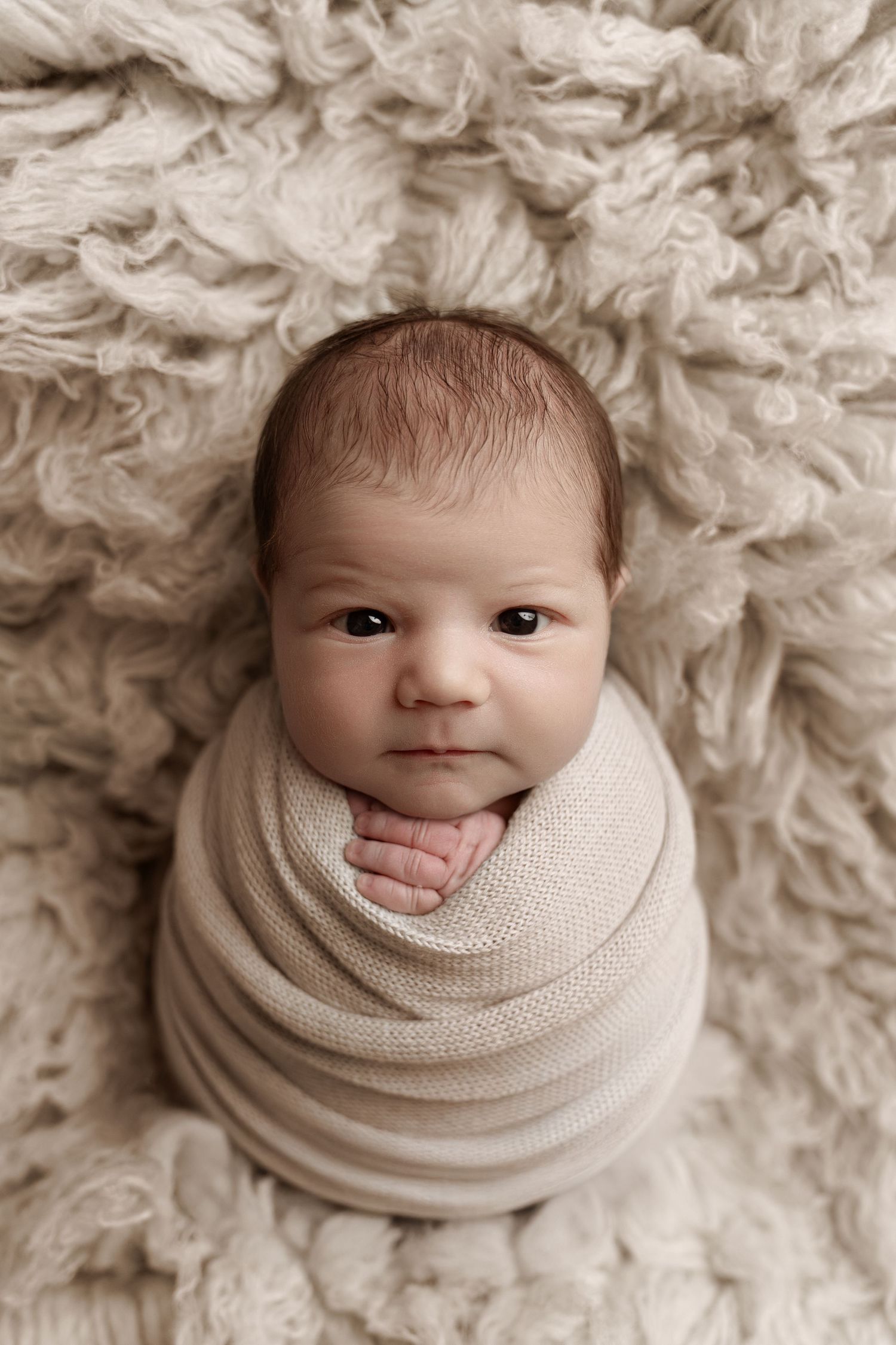 A swaddled newborn baby wrapped in a cream knit blanket lies on a fluffy beige rug, looking up with dark eyes.