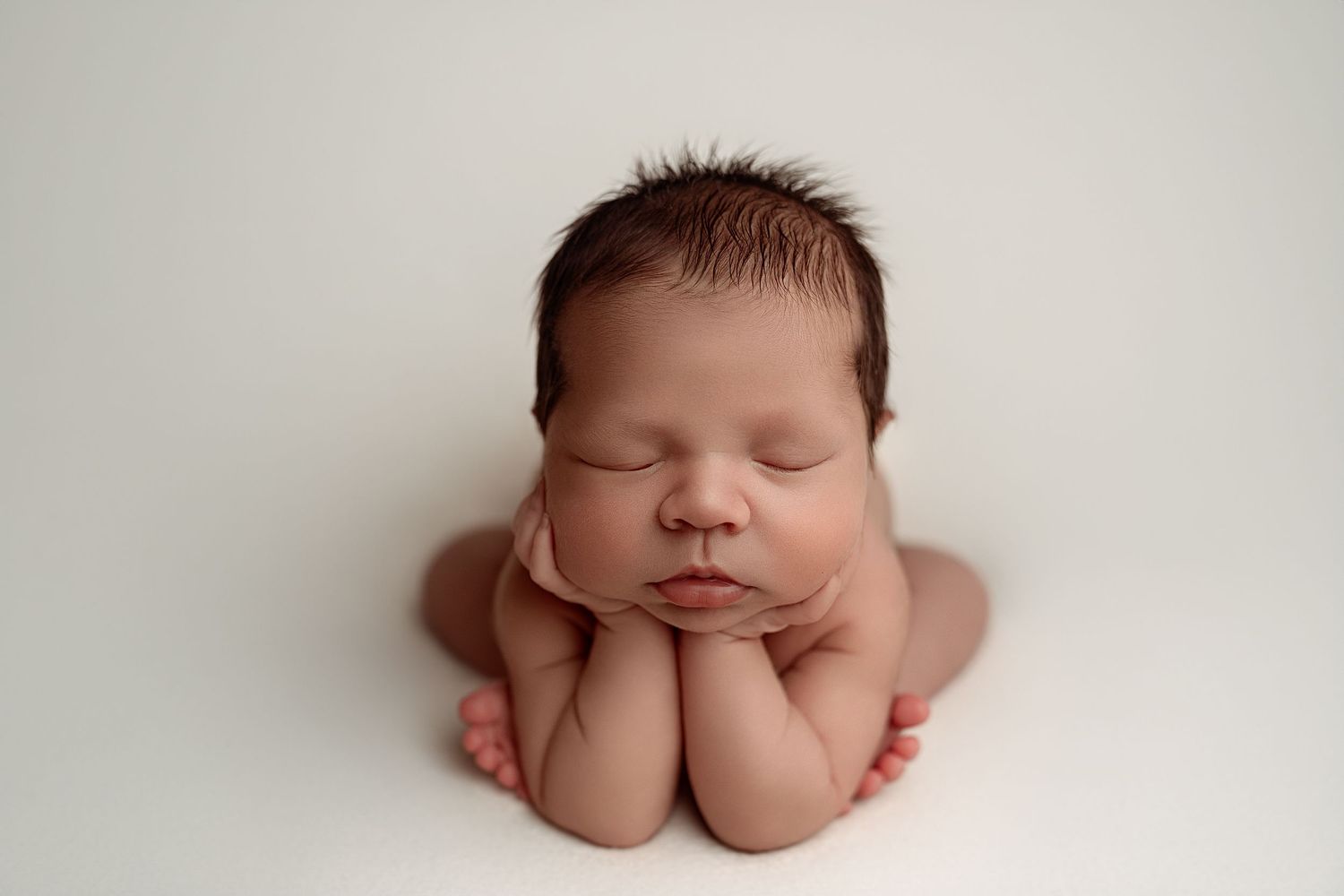 Peaceful sleeping newborn baby with dark hair resting chin on hands in classic froggy pose on white background.