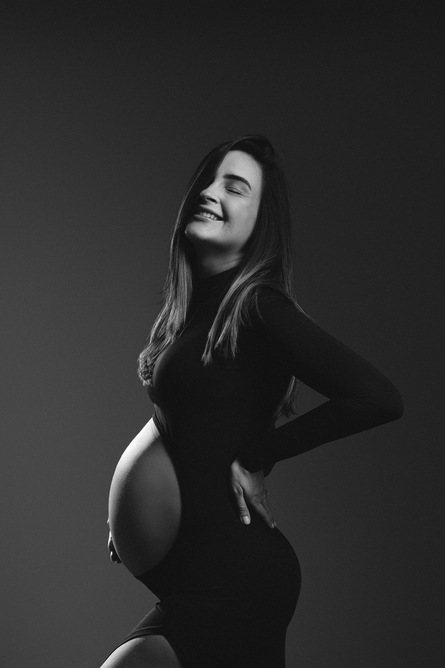 Black and white portrait of a smiling pregnant woman in a dark bodysuit cradling her baby bump against a dark background.