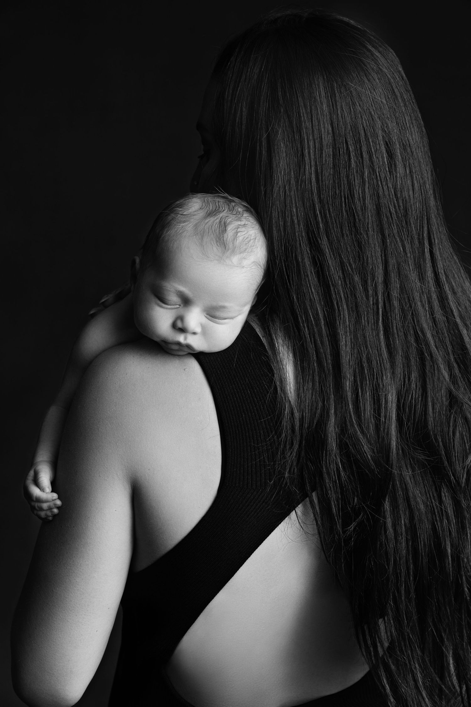 Black and white photo of a mother holding a sleeping newborn baby against her shoulder, dark background.