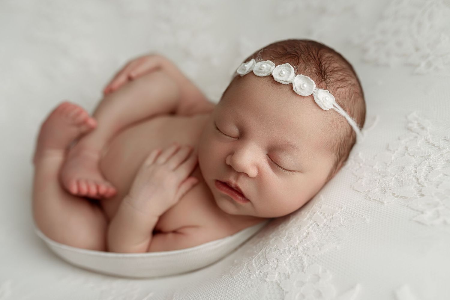 Peaceful newborn baby girl sleeping curled in white bowl, wearing delicate floral headband, on soft lace backdrop.