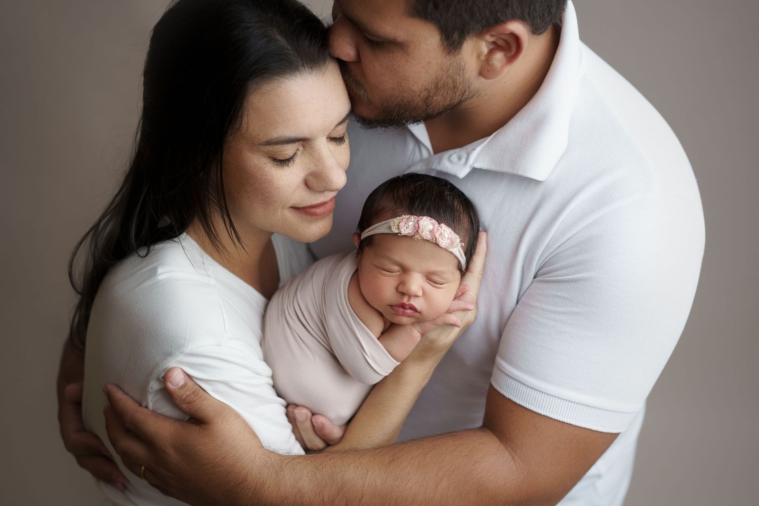 Parents tenderly hold their sleeping newborn baby girl wearing a pink floral headband in a professional studio portrait.