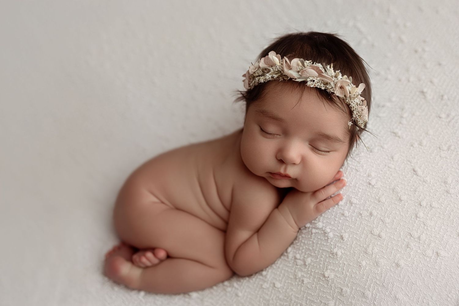 Peaceful sleeping newborn baby girl wearing a delicate floral headband, posed on a soft white textured blanket.