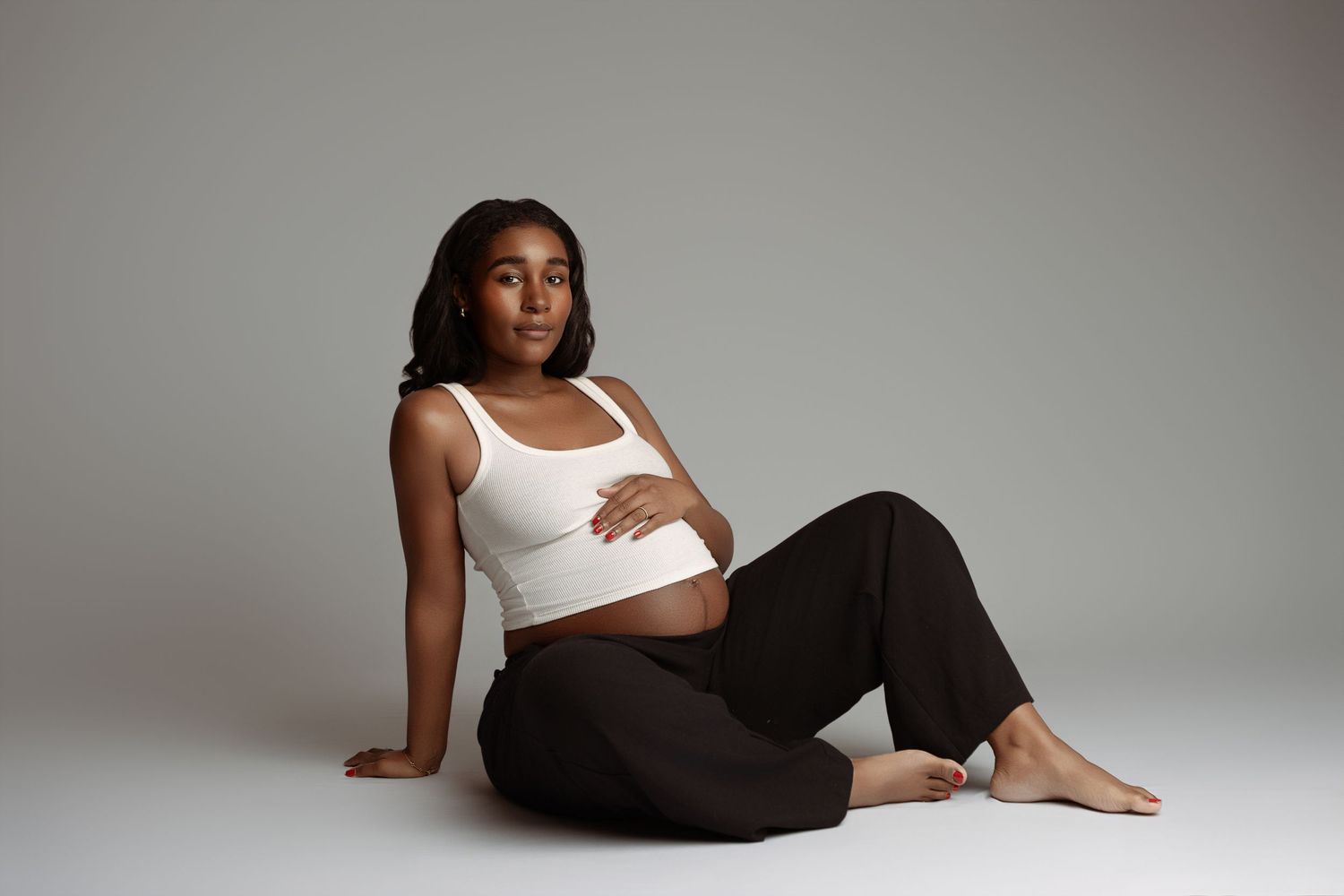 Pregnant woman in white crop top and black pants sitting on floor, cradling baby bump in studio setting.
