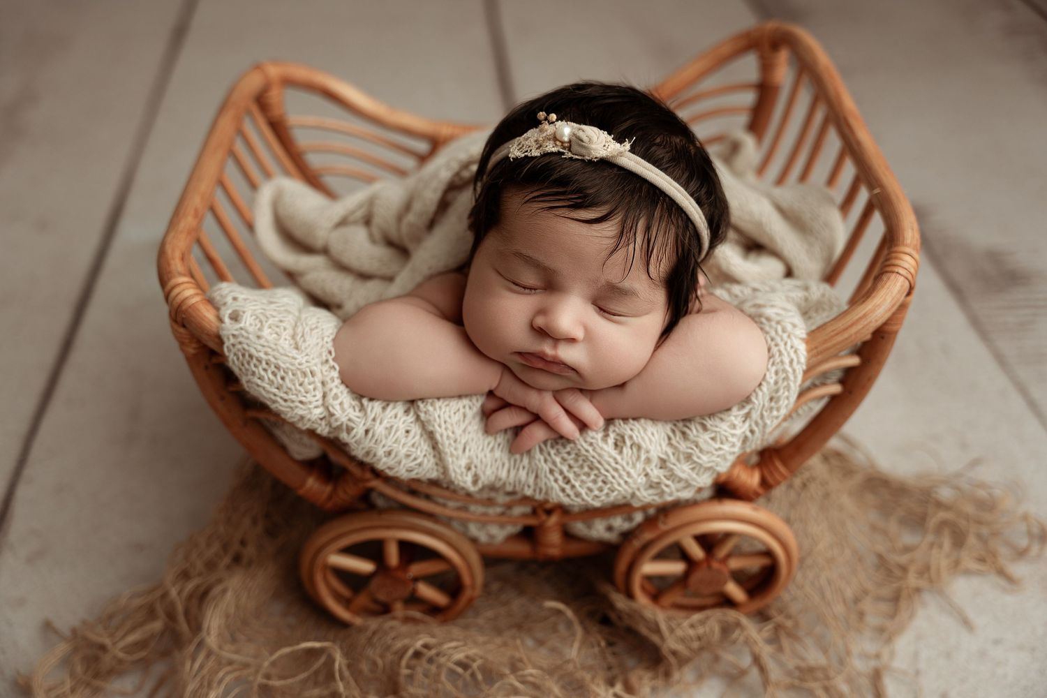 Newborn baby sleeping peacefully in a wicker carriage prop with cream knit wrap and floral headband.