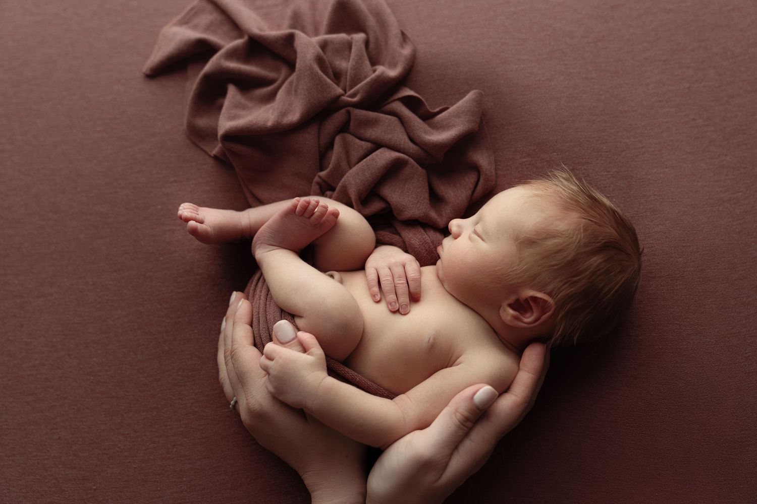 Newborn baby sleeping peacefully in adult hands on a brown fabric background.