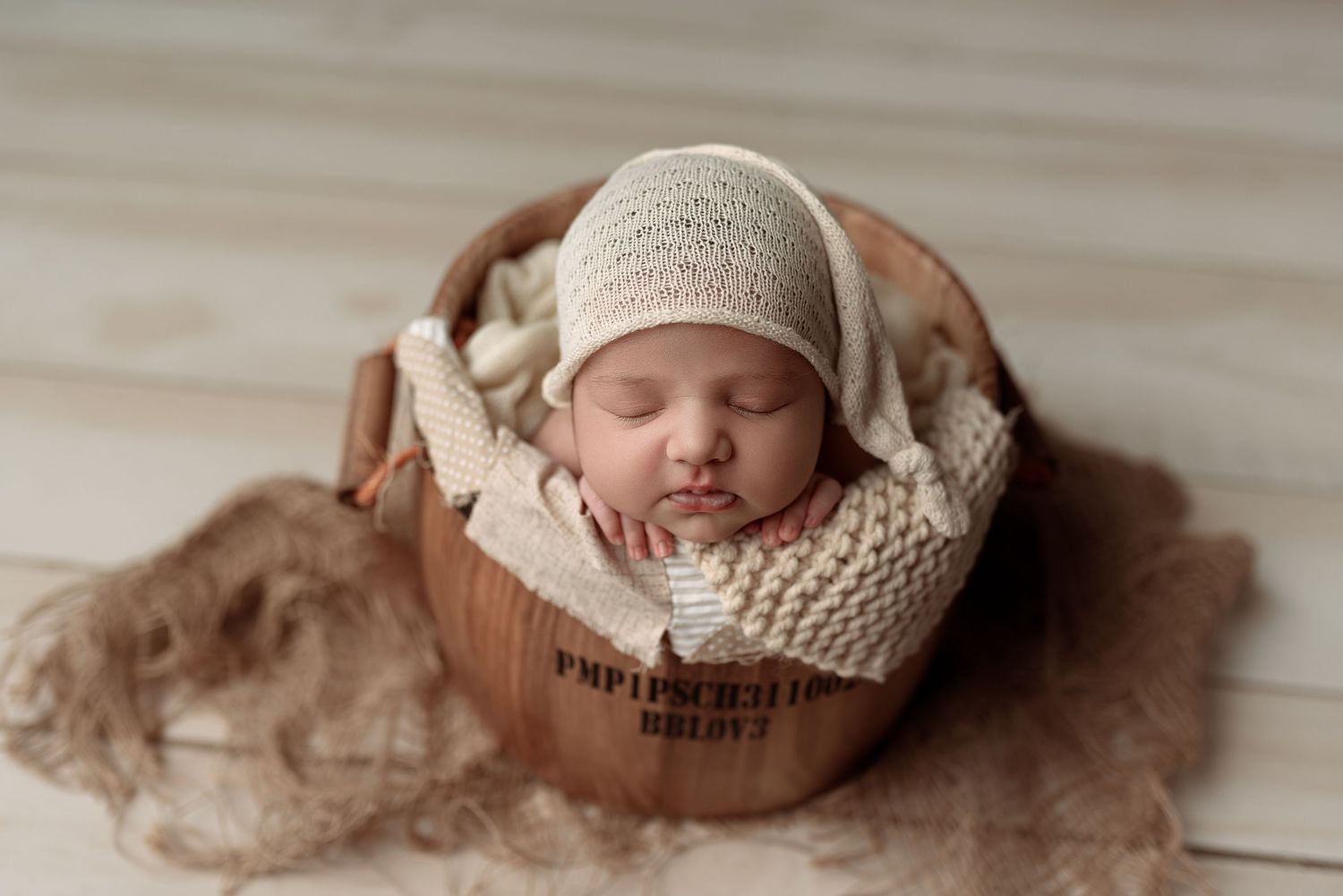 Sleeping newborn baby in cream knit bonnet nestled in wooden bucket with soft beige wraps on light wood floor.