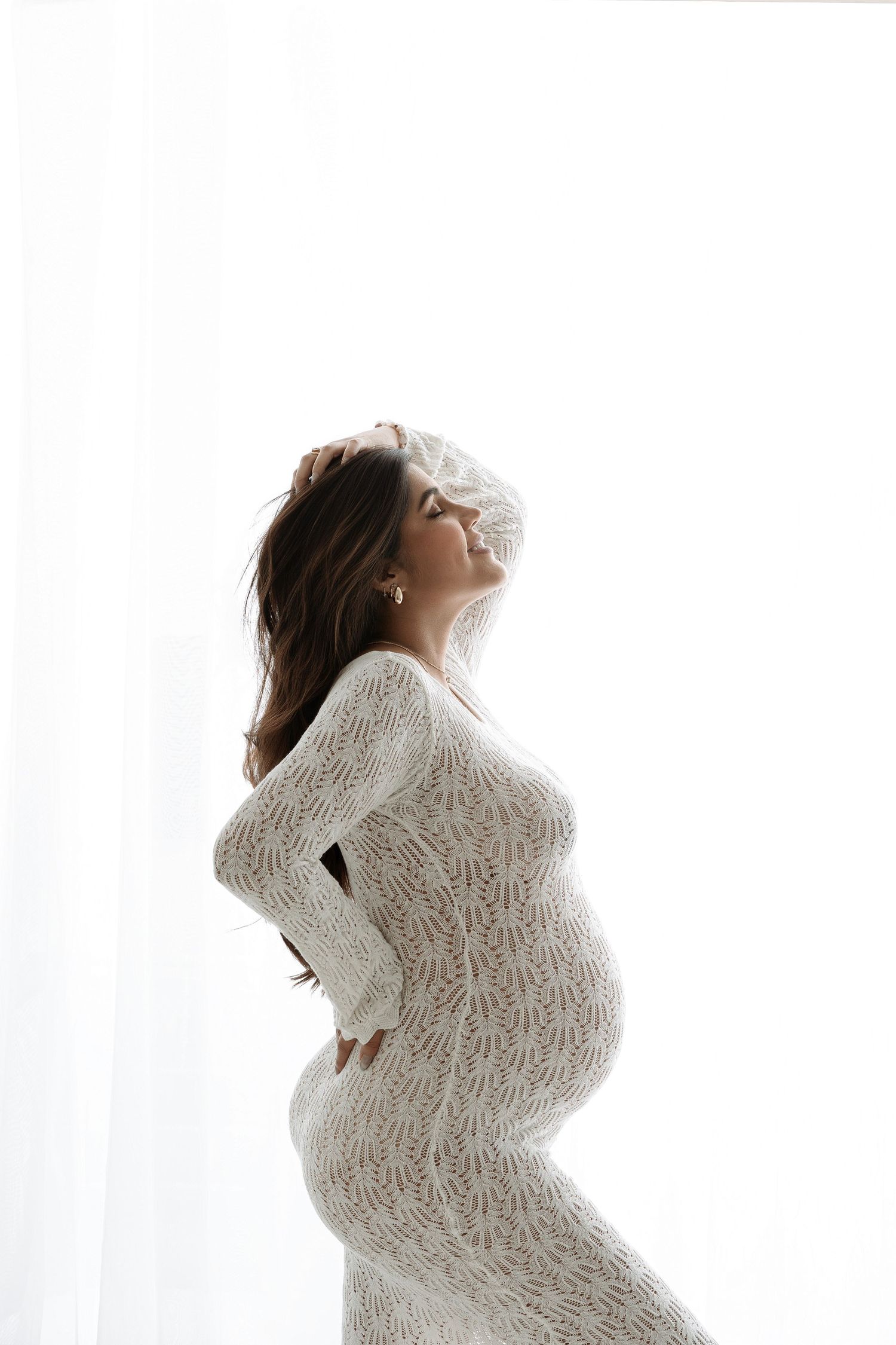 Pregnant woman in white lace dress standing by bright window, hands on back, glowing in natural light.