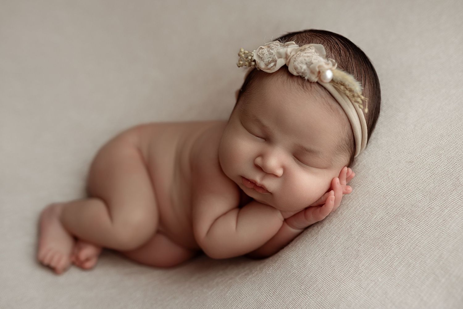 Peaceful sleeping newborn baby girl wearing a delicate floral headband, posed on a soft beige blanket.