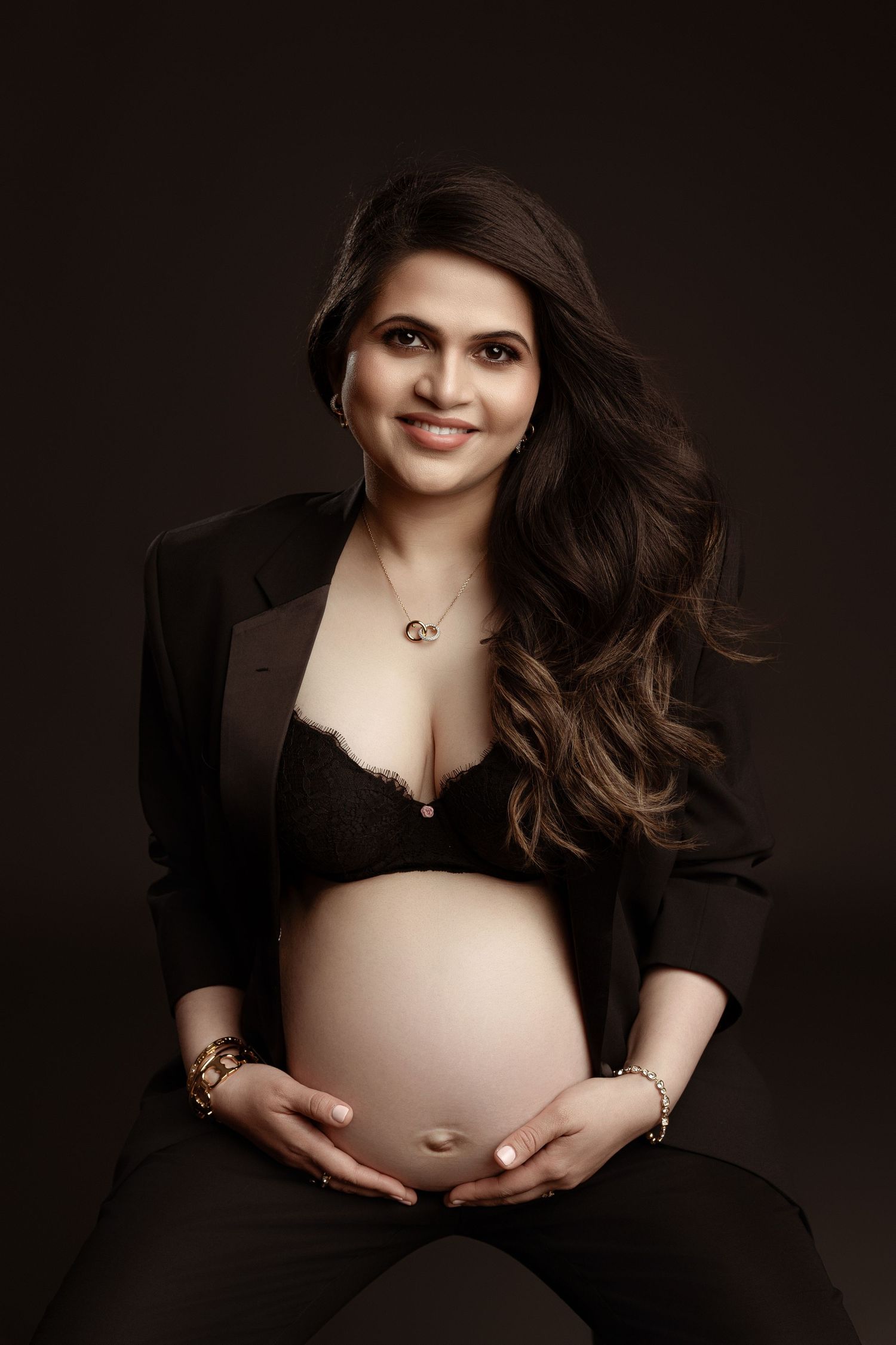 Smiling pregnant woman in black blazer and lace bralette cradling baby bump in studio portrait.