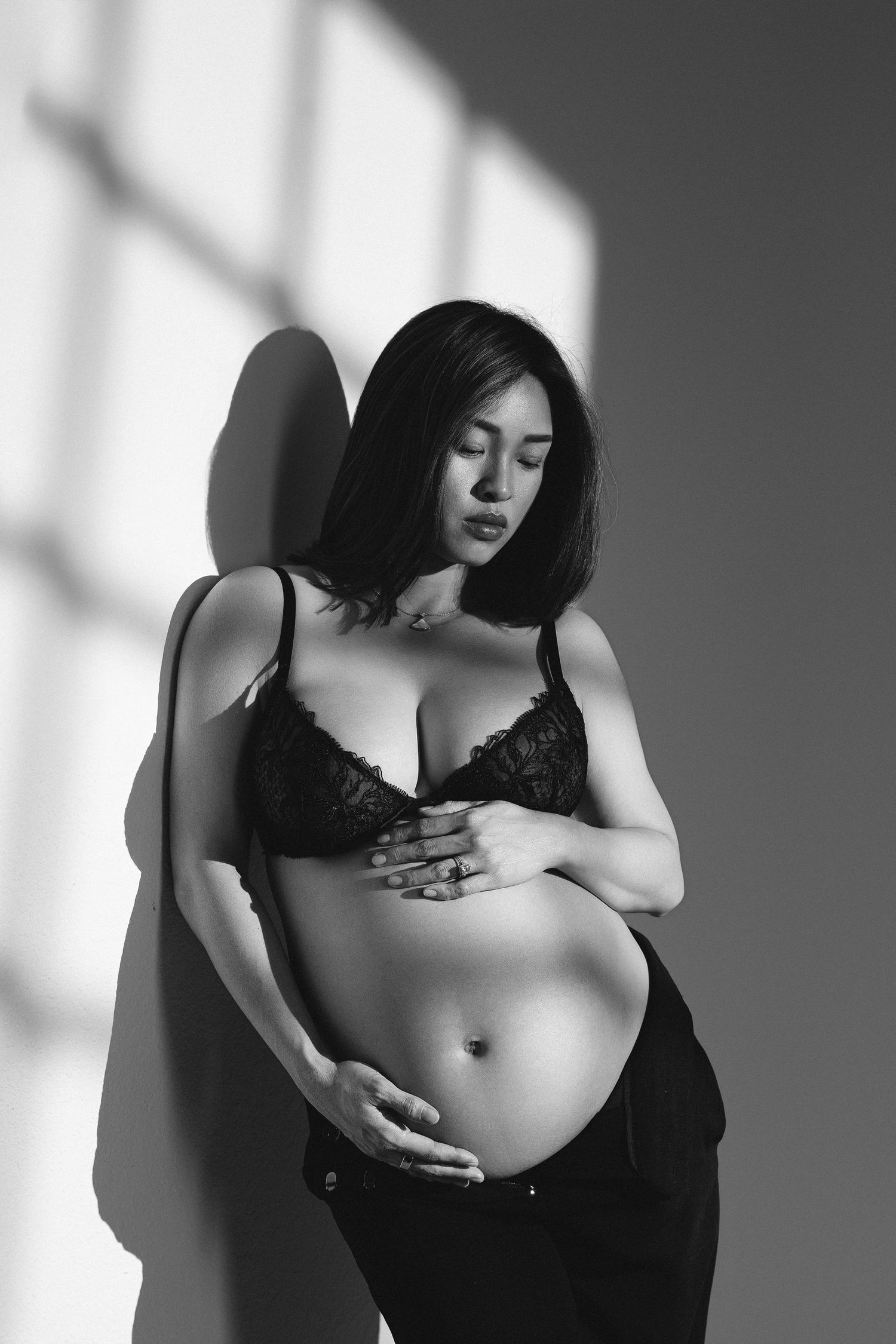 Black and white photo of a pregnant woman in lace bra cradling her baby bump against a sunlit wall.