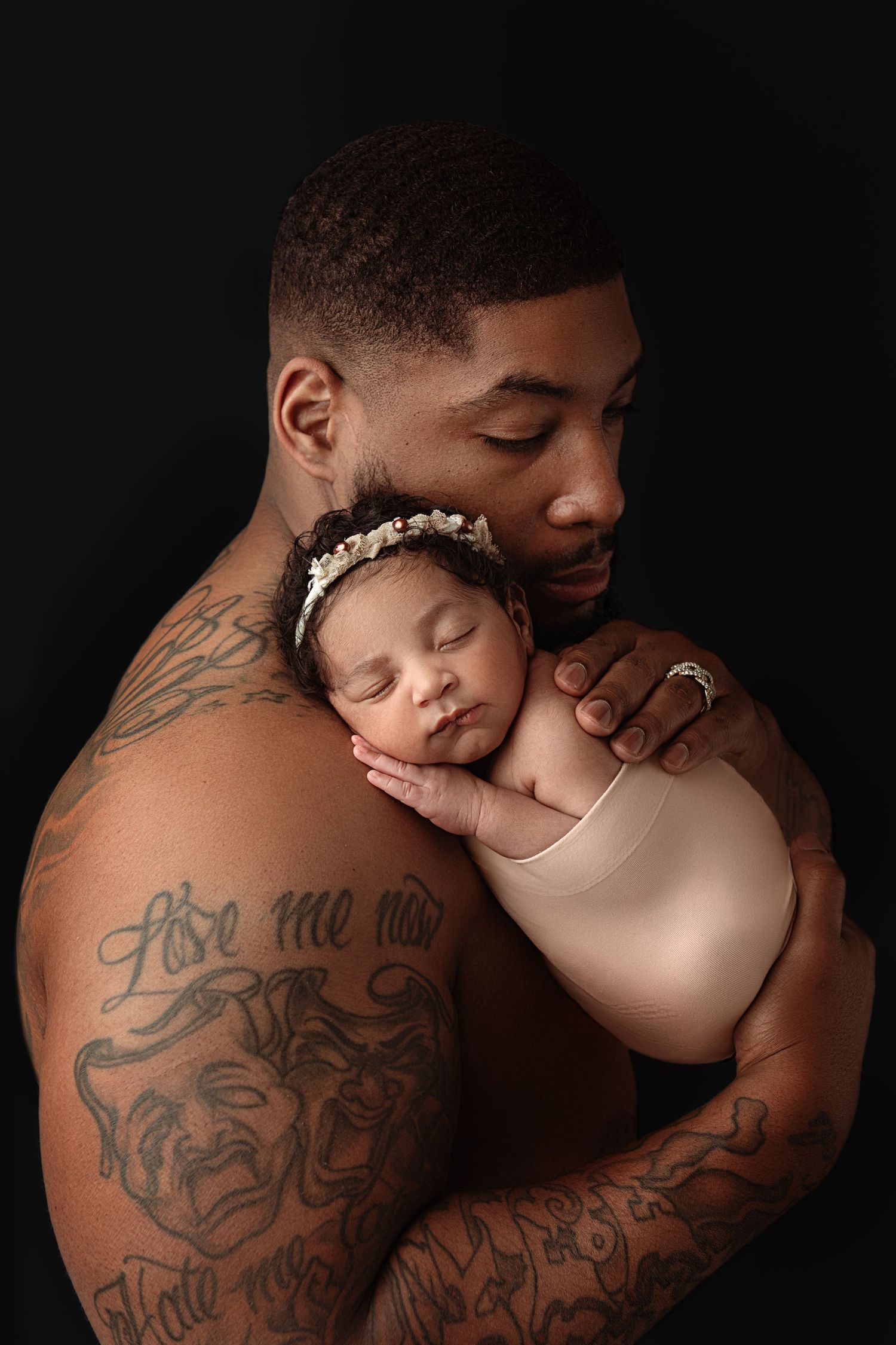 A tender moment captured as a tattooed parent holds their sleeping newborn baby against their chest in a dark studio portrait. Paloma Schell