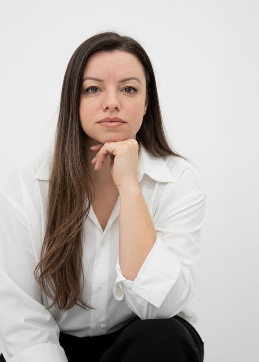 Professional woman in white shirt with long dark hair poses thoughtfully with hand on chin against white background.