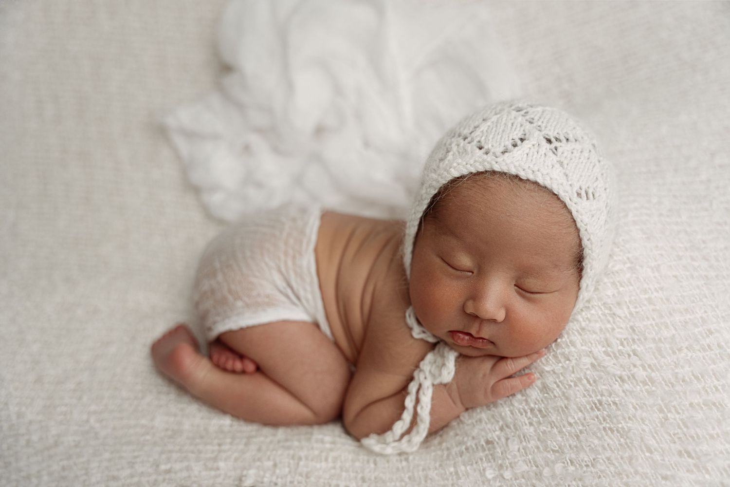 Sleeping newborn baby in white knit bonnet and romper posed on soft white blanket.