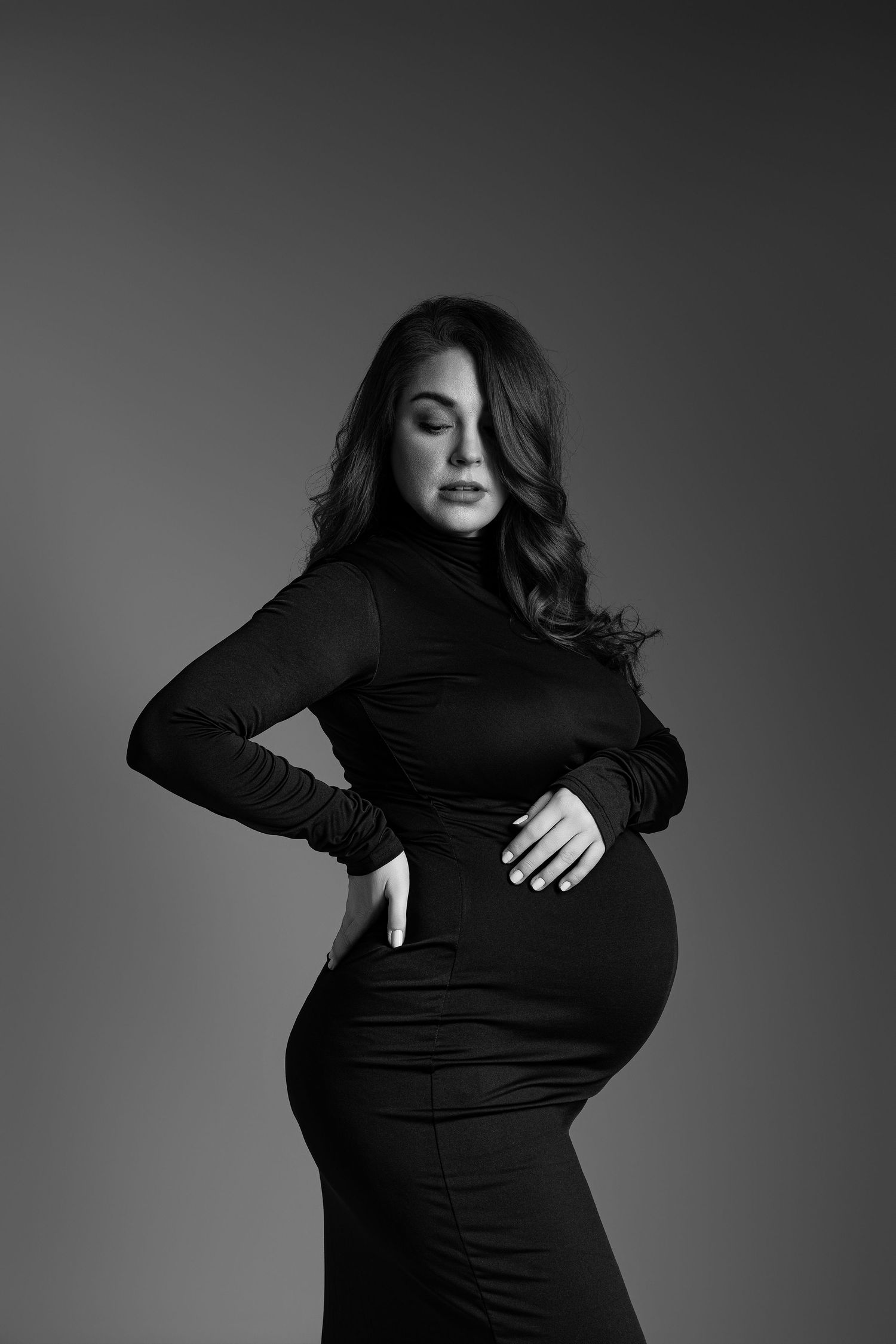 Black and white maternity photo of a pregnant woman in a black turtleneck dress cradling her baby bump.