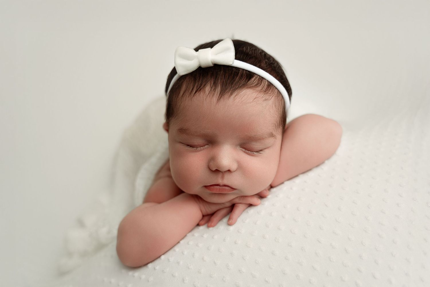 Sleeping newborn baby girl wearing a white bow headband, resting on a soft white blanket in a studio portrait.
