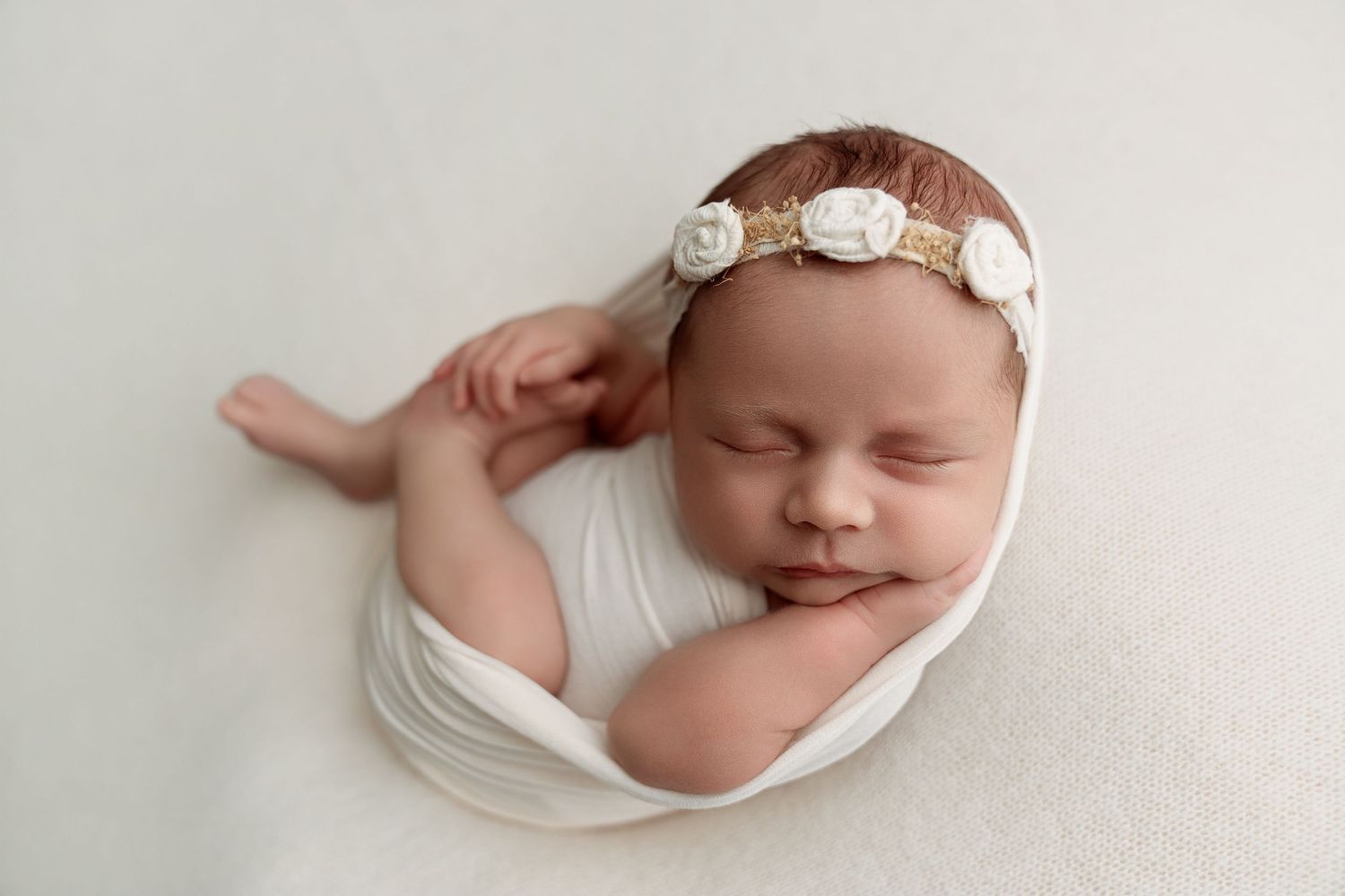 Sleeping newborn baby girl wrapped in white swaddle with floral headband on soft white backdrop.