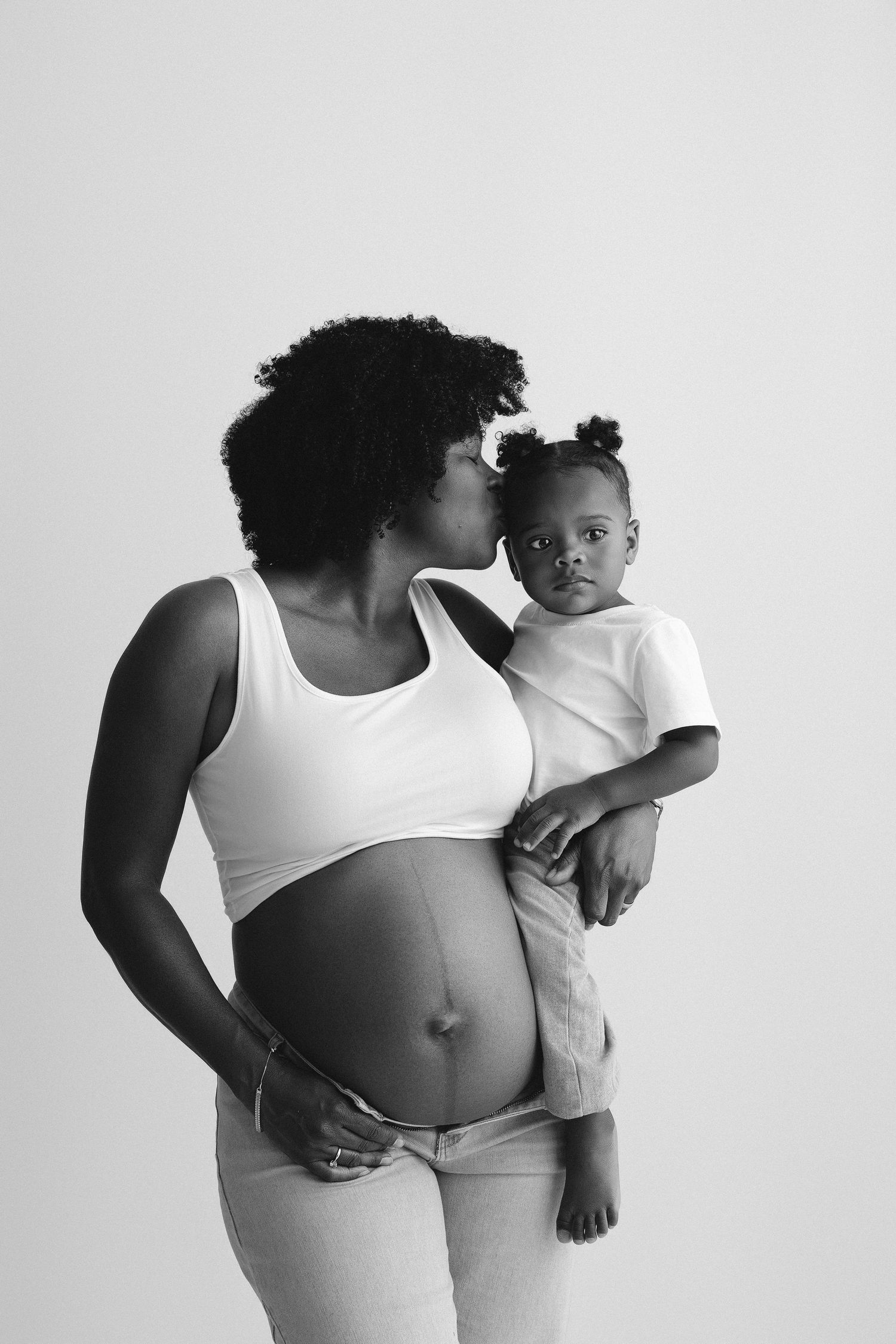 Black and white photo of a pregnant woman kissing her toddler while holding them, showcasing a beautiful maternity moment.