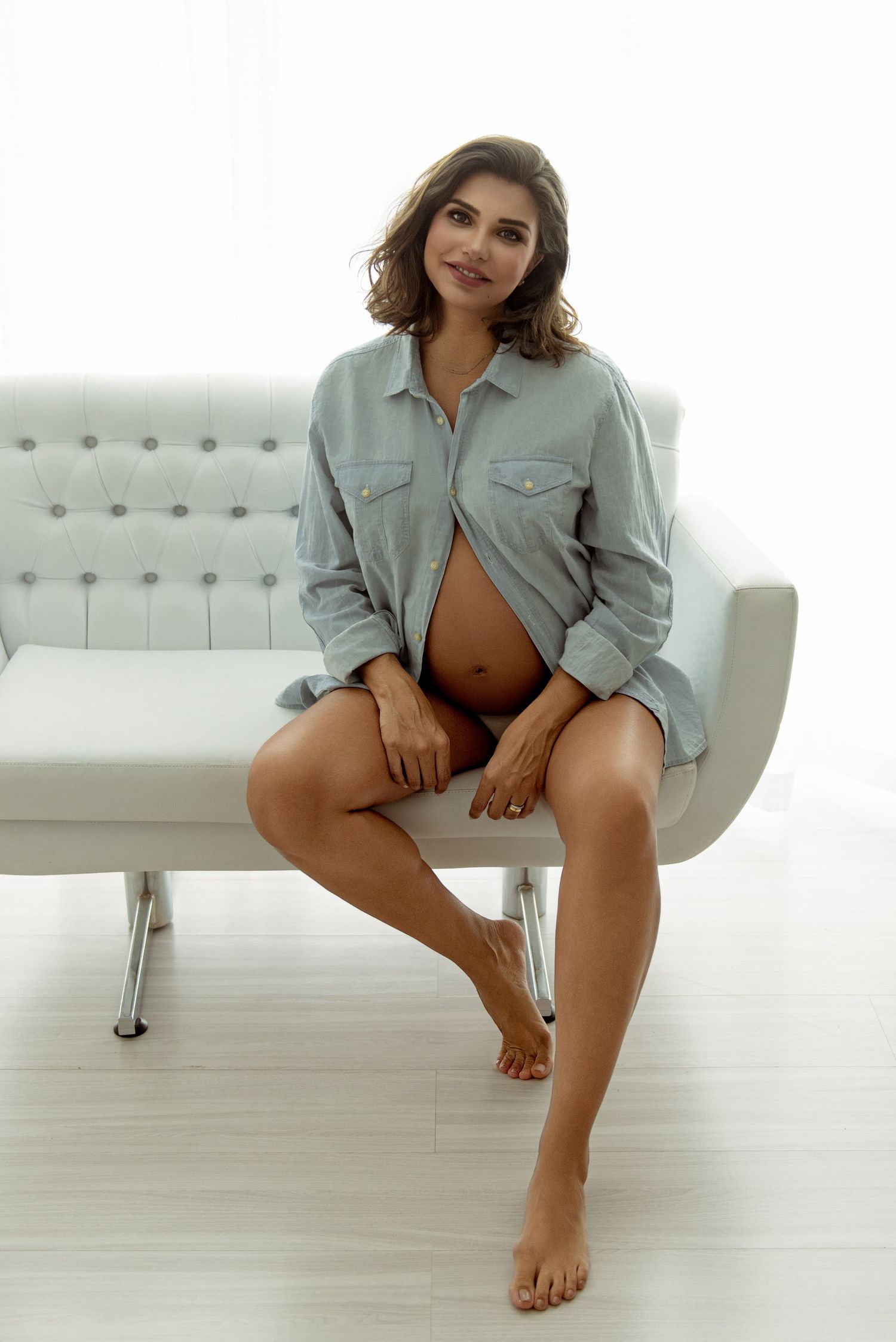 Pregnant woman in open denim shirt sitting barefoot on white tufted sofa, showing baby bump in bright minimalist room.