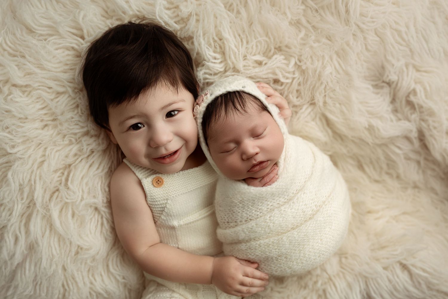 Two young children laying together on a soft white fur rug in matching cream-colored outfits– newborn photographer Paloma Schell Houston TX
