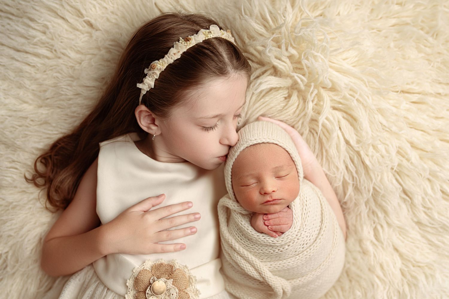 Older sister in white dress kissing sleeping newborn baby wrapped in cream swaddle on fluffy white blanket.