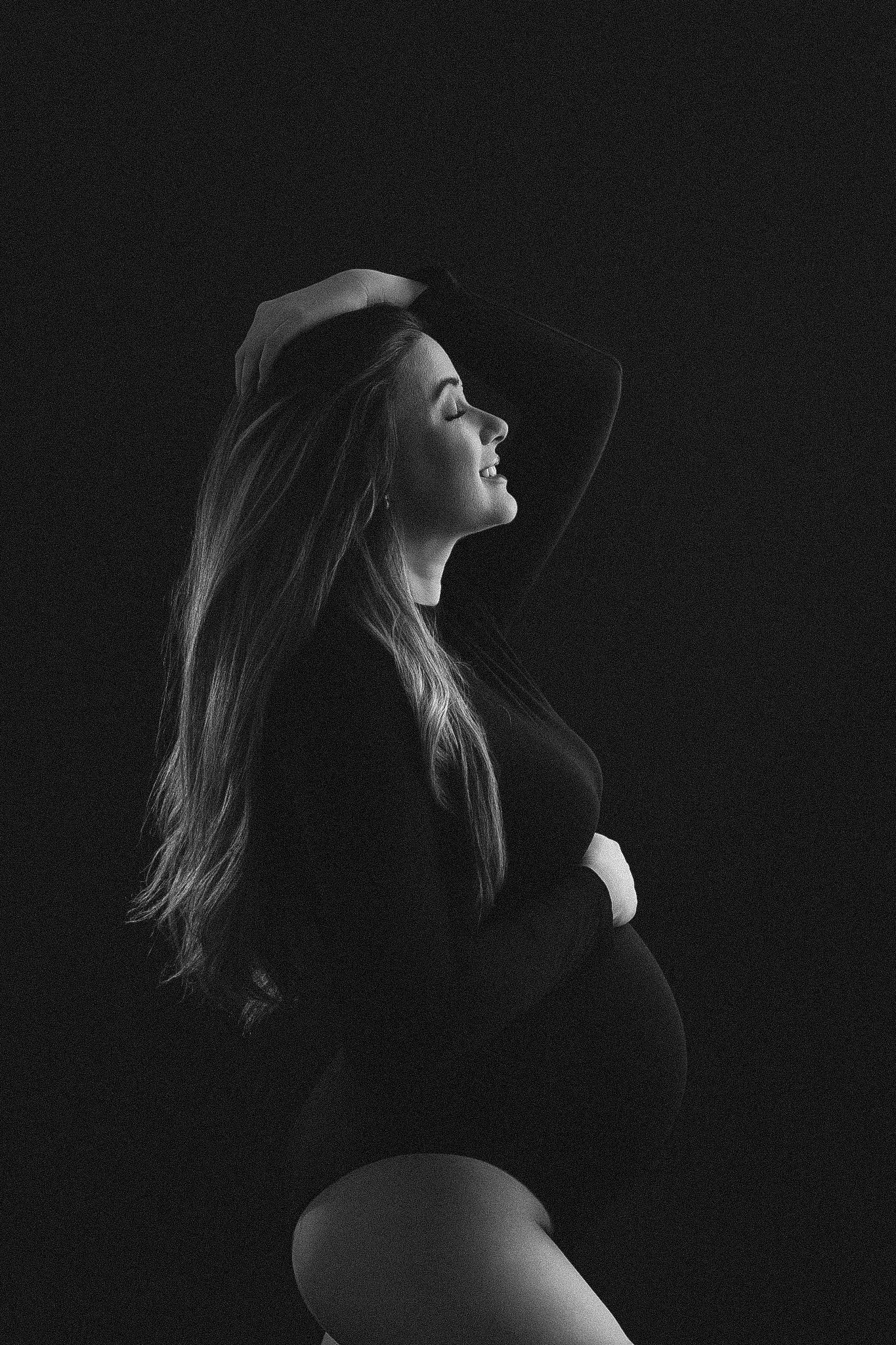 Black and white maternity portrait of a pregnant woman cradling her belly, posing gracefully against a dark background.