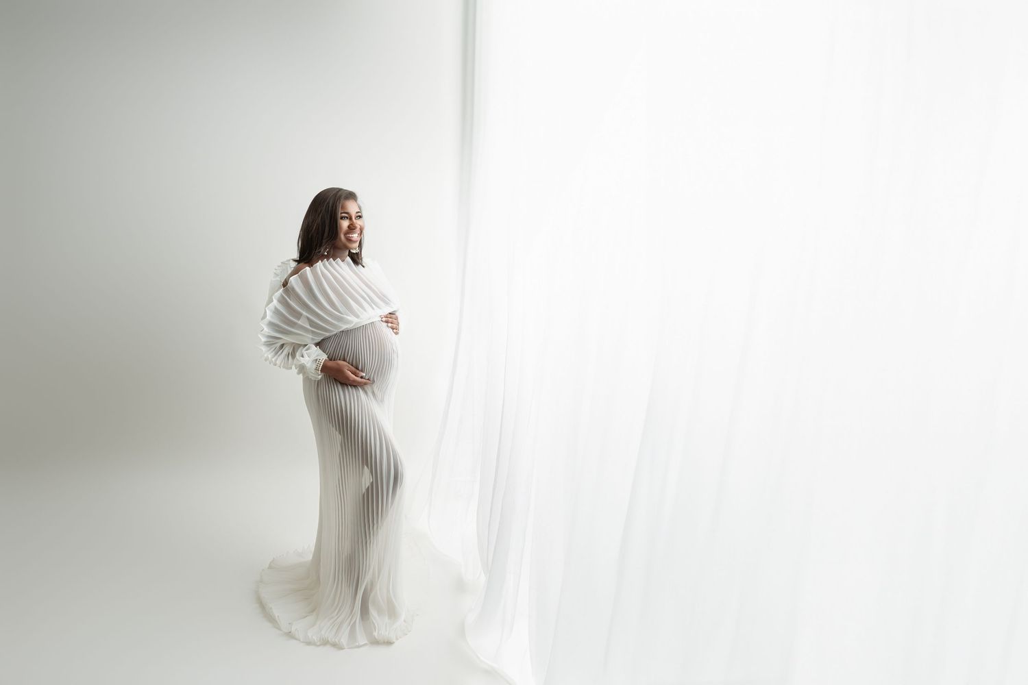 Pregnant woman in white ruffle gown holding baby bump beside flowing white curtain in bright studio.