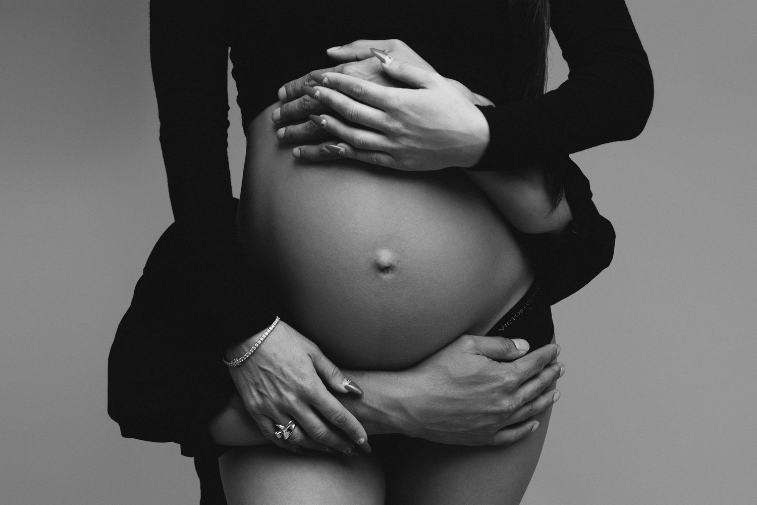 Black and white maternity photo of a pregnant woman's bare belly being cradled by two pairs of hands.