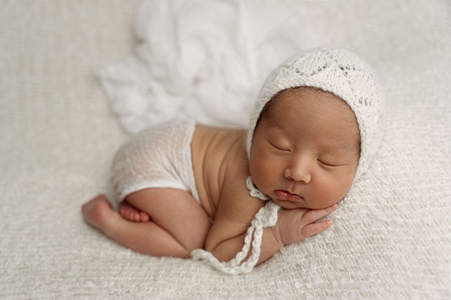 A sleeping newborn baby wearing white lace clothing lies peacefully on soft white textured fabric. – newborn photographer Paloma Schell